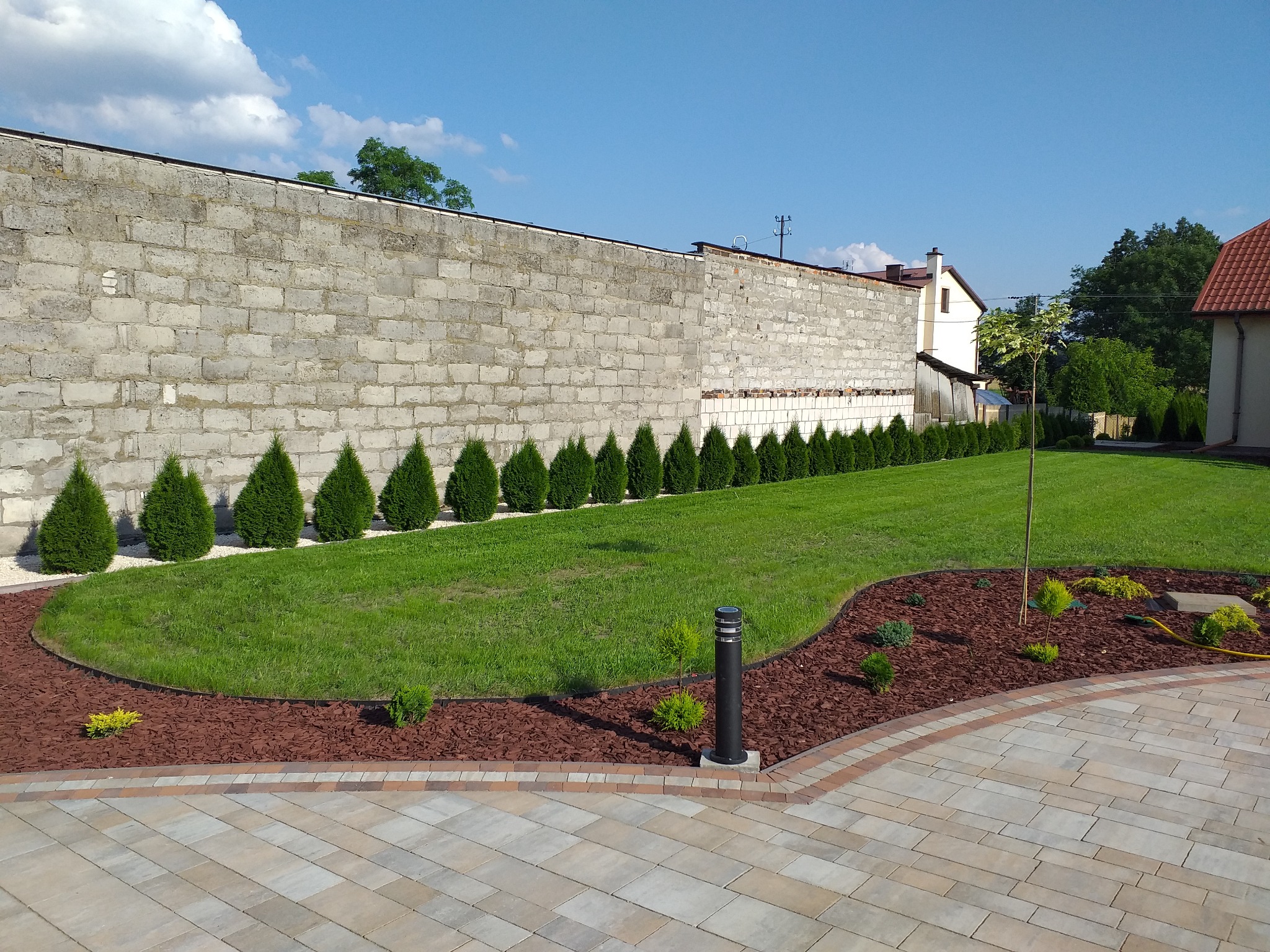 Uporządkowany ogród z równo przyciętym trawnikiem, rządkiem stożkowych iglaków wzdłuż szarego muru z bloczków, rabatą z brązową korą i niskimi krzewami, oraz chodnikiem z kostki brukowej...