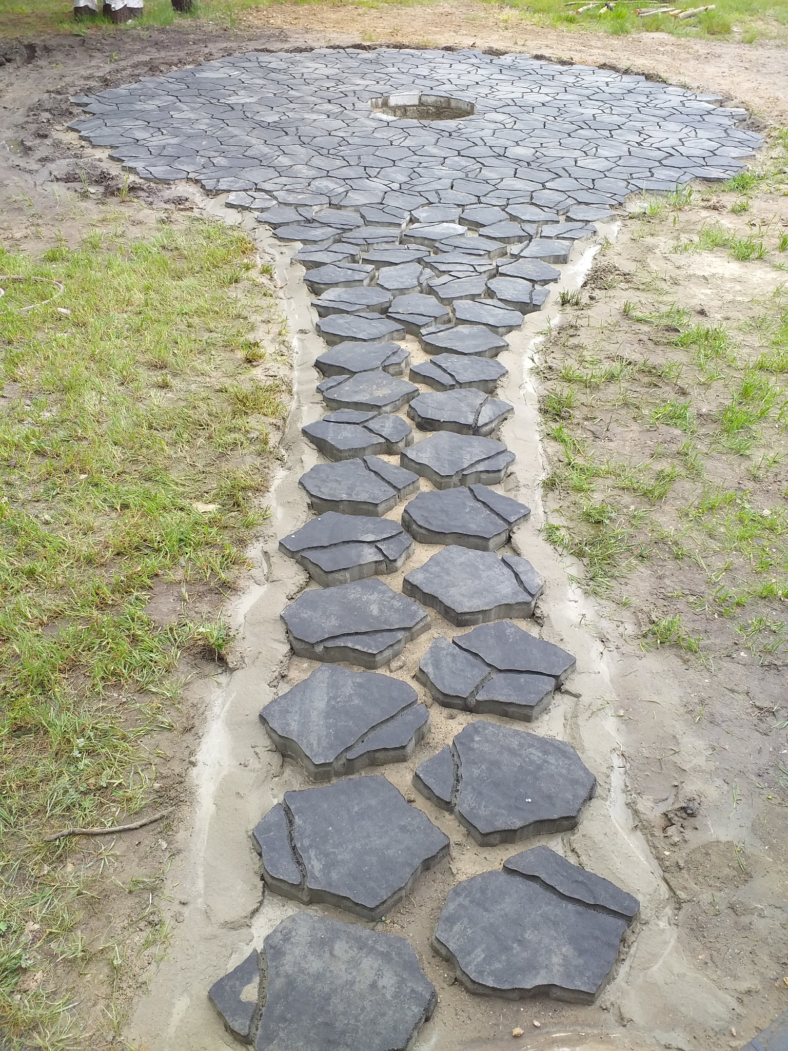 Układanie ścieżki z nieregularnych, szarych płyt betonowych w ogrodzie, widoczny piasek i trawa, w tle okrągły plac z podobnej kostki z otworem pośrodku.