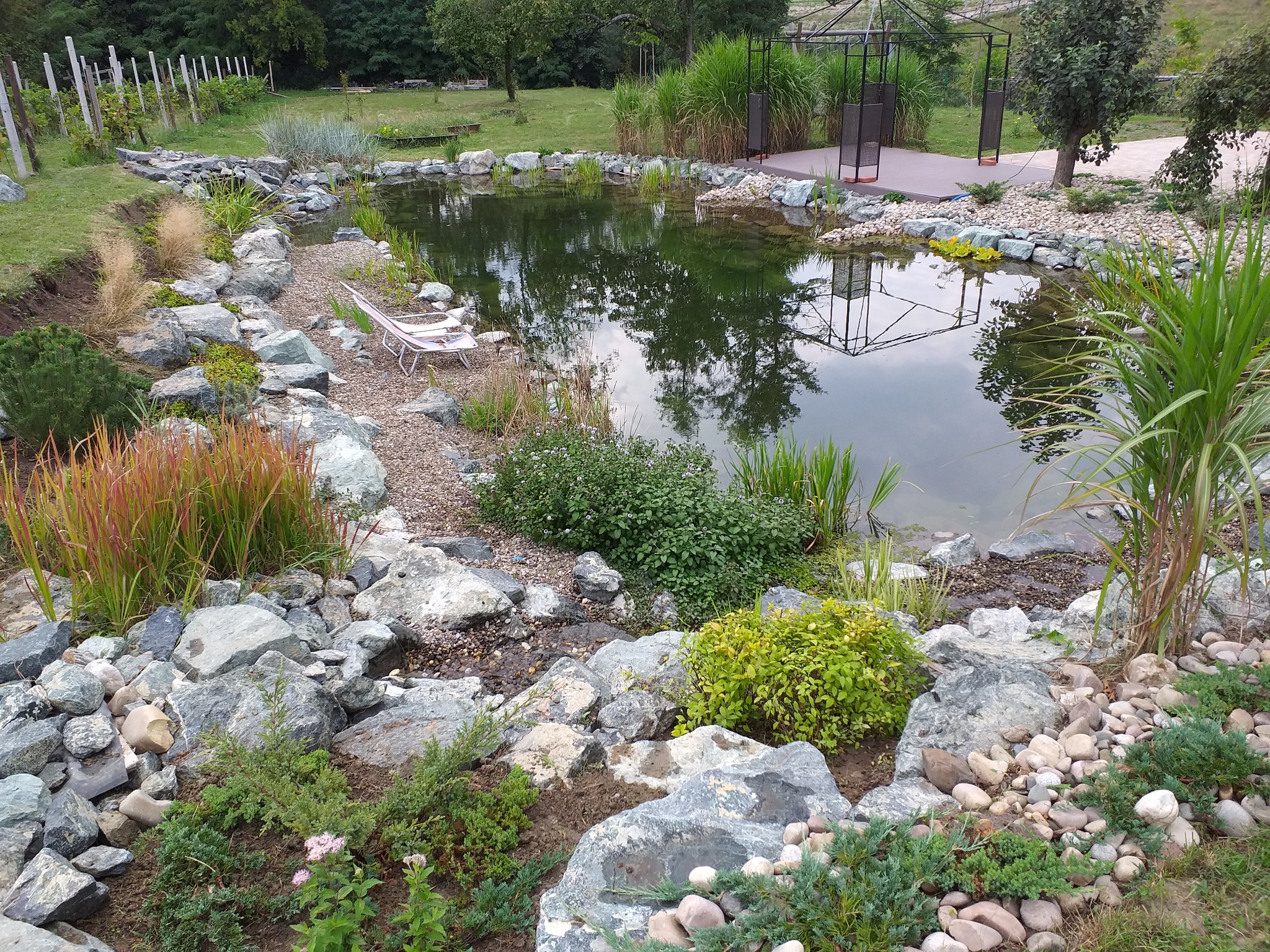 Oczko wodne z kamiennym brzegiem, roślinnością ozdobną, trzcinami i leżakiem, odbicie altany w tafli wody.