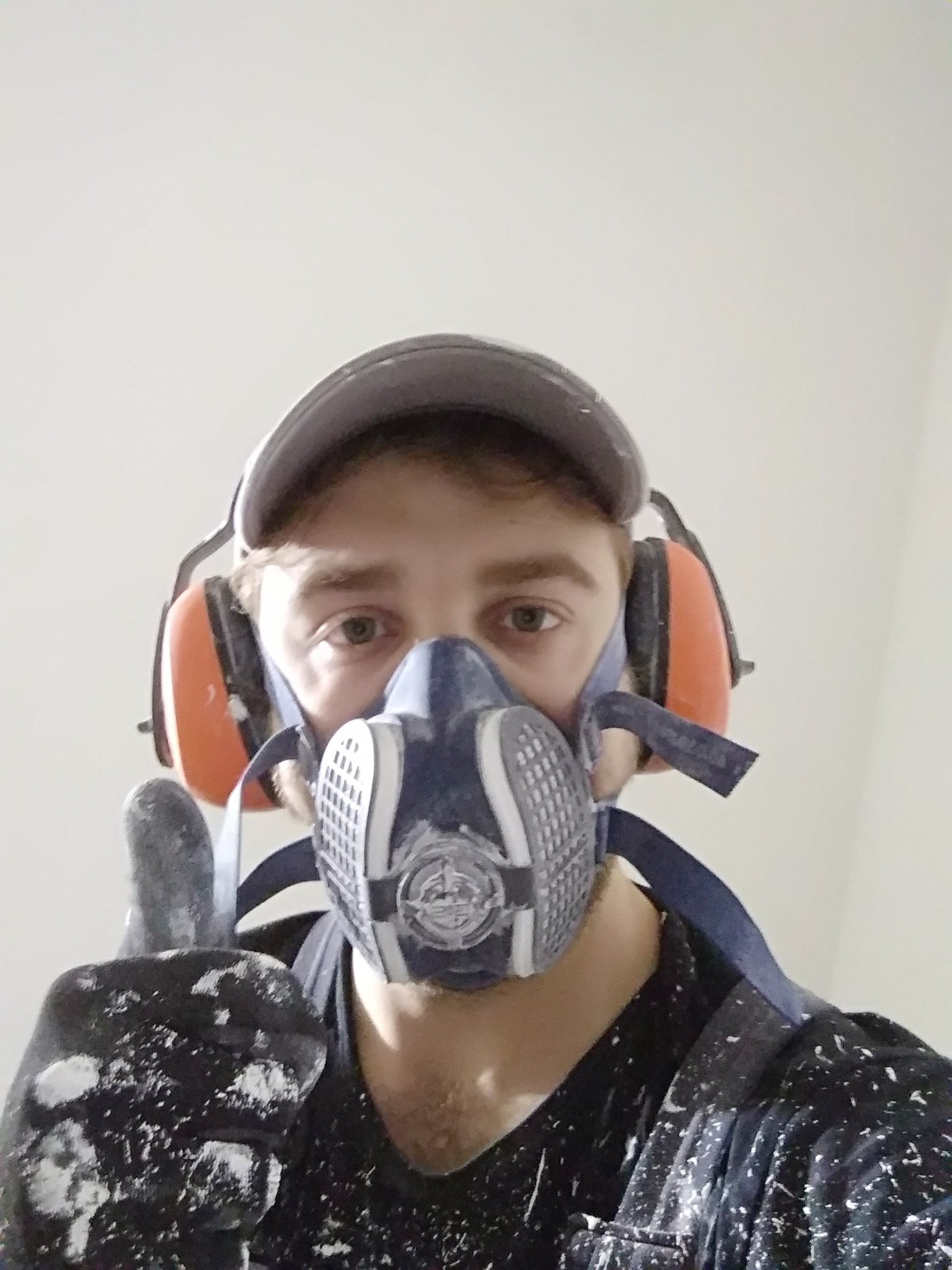 Mężczyzna w czapce, słuchawkach ochronnych i masce przeciwpyłowej, ubrudzony farbą, pokazuje kciuk w górę, prawdopodobnie po malowaniu ścian.