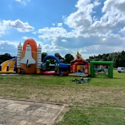 Kompleks dmuchańców na trawie: rakieta kosmiczna, tor przeszkód, klaun z basenem, bramka piłkarska, w tle samochód strażacki i dostawczy.