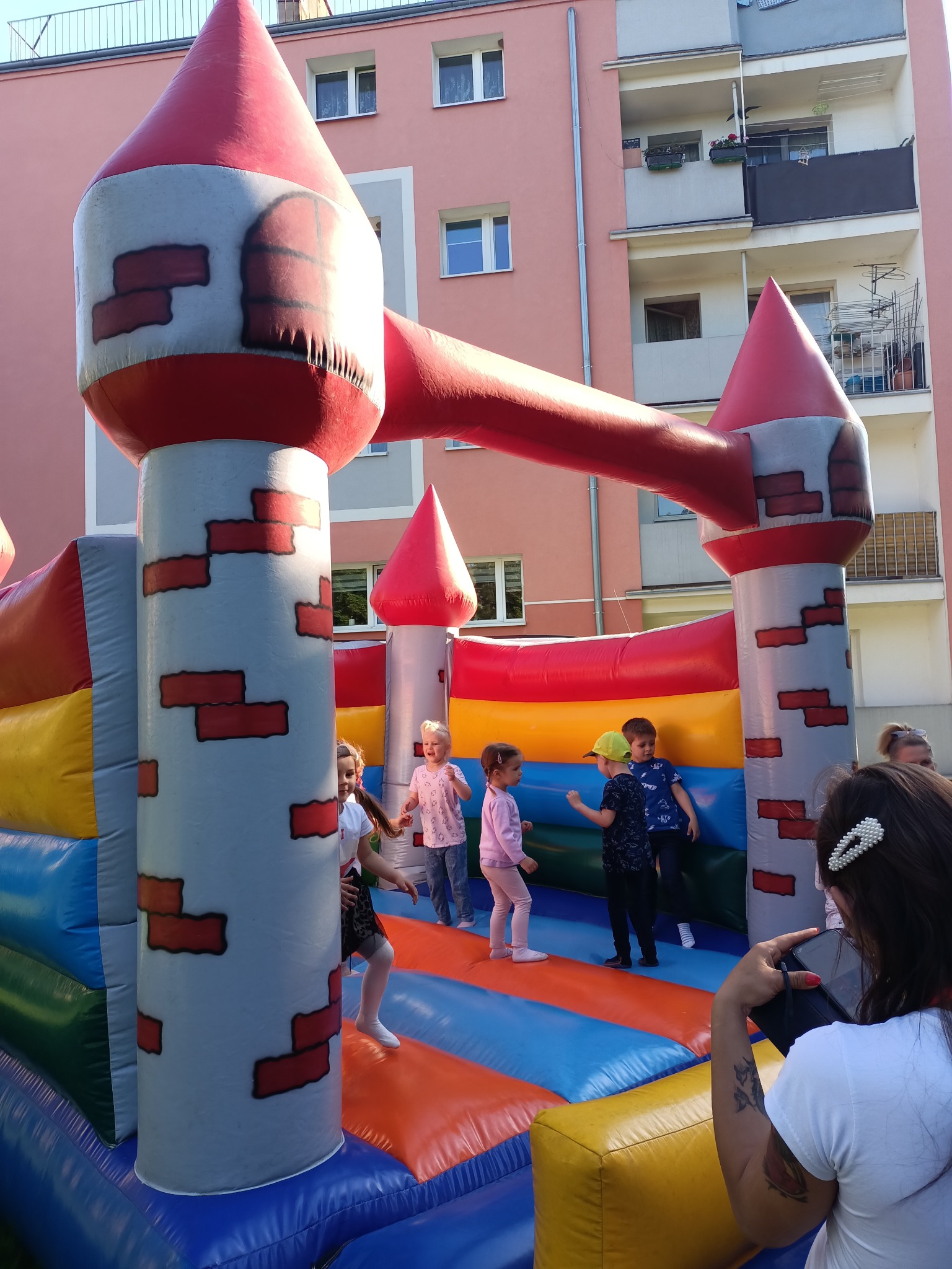 Dmuchany zamek w kształcie średniowiecznej fortecy z czerwonymi wieżami i szarymi murami, na którym bawi się grupa dzieci w różnym wieku, na tle bloku mieszkalnego. Kobieta z tatuażem na ramieniu...