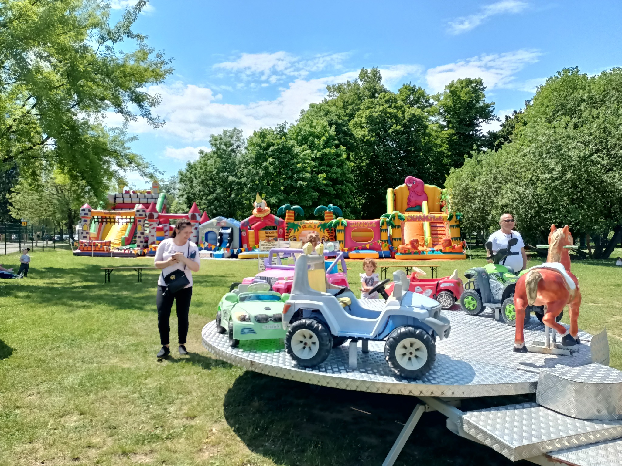 Kolorowe dmuchańce i karuzela z elektrycznymi autkami na trawiastym terenie, dzieci korzystające z atrakcji, widok w słoneczny dzień.