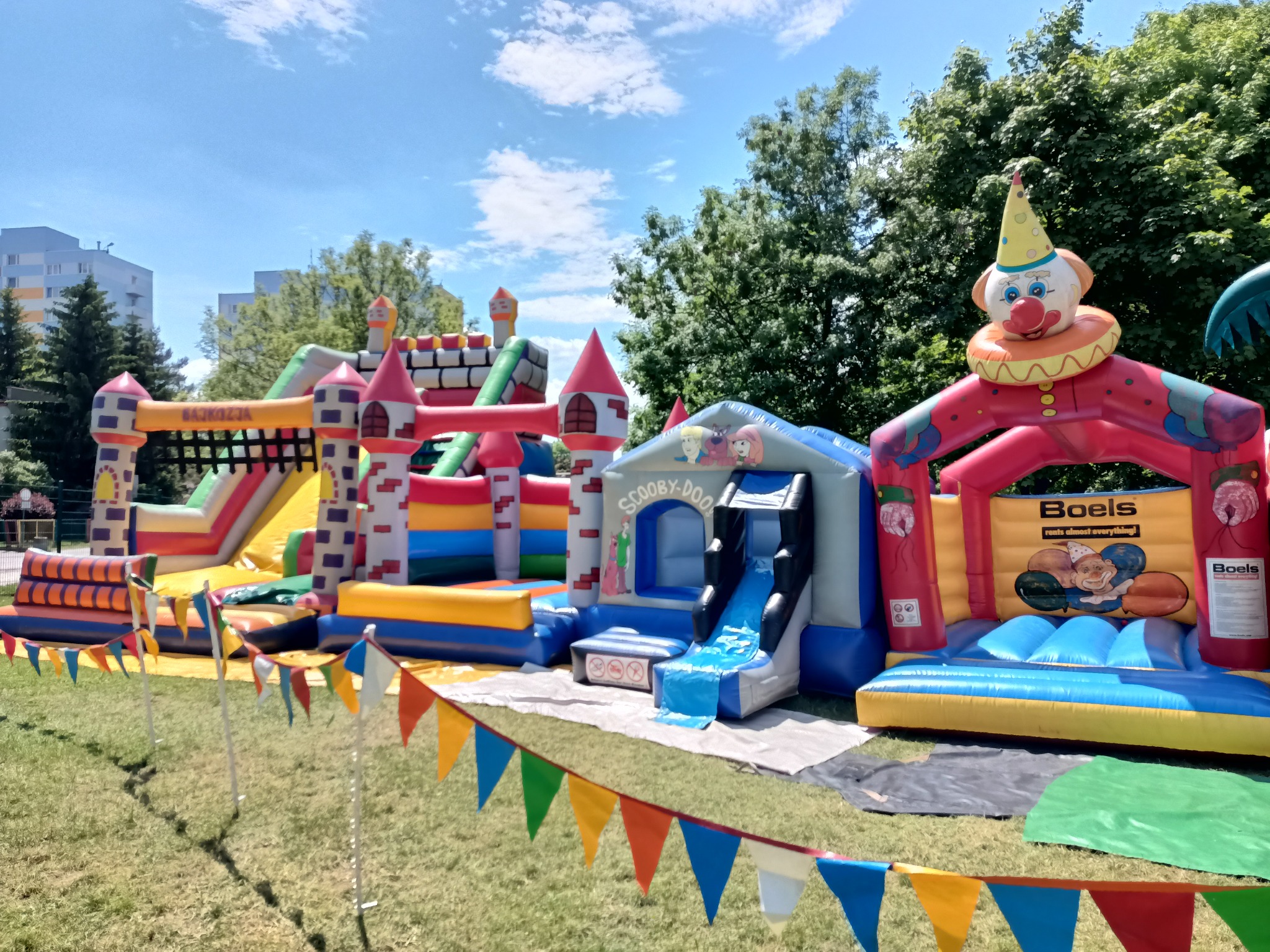 Kompleks dmuchańców na trawie z dekoracyjnymi chorągiewkami, obejmujący zamek ze zjeżdżalnią, dmuchany plac zabaw z motywem Scooby-Doo i dmuchany zamek z klaunem w koronie.
