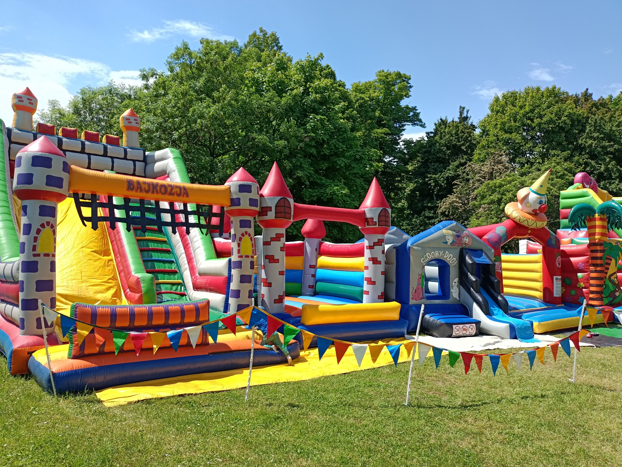 Kompleks dmuchanych zamków i zjeżdżalni w jaskrawych kolorach, otoczony zielonym trawnikiem, z dekoracyjnymi girlandami z trójkątnych flag.