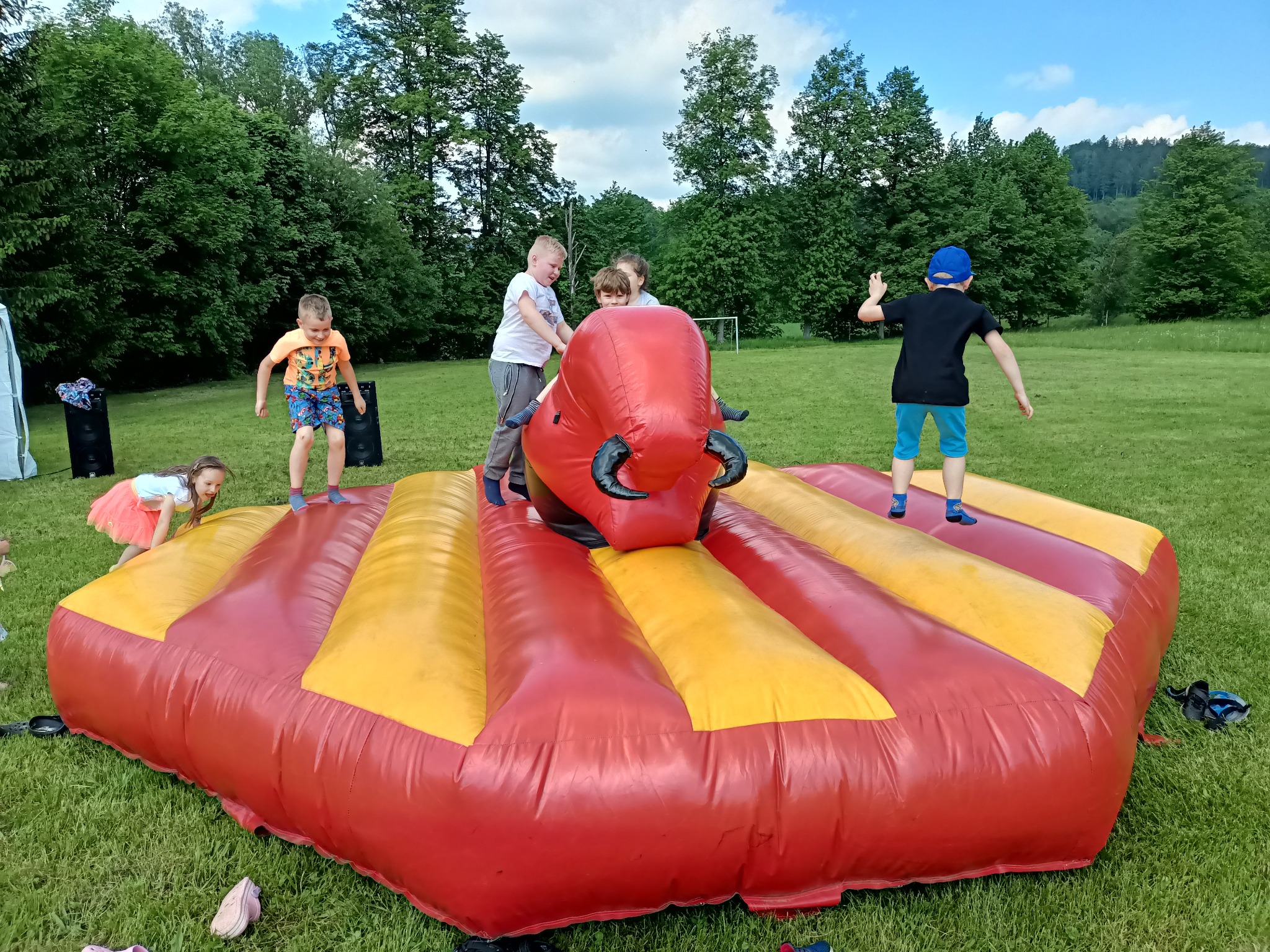 Dzieci bawią się na dmuchanym byku na trawiastym terenie z drzewami w tle, widać głośniki i buty zdjęte przez dzieci.