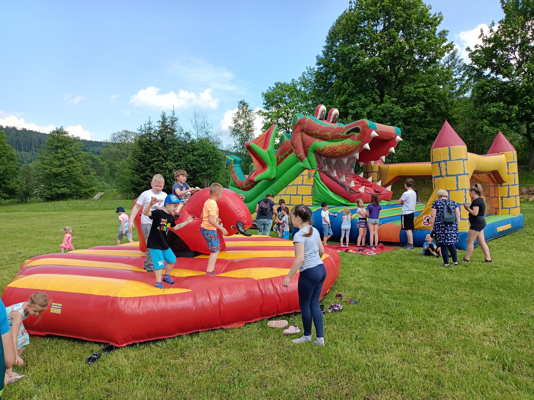 Dzieci bawią się na dmuchanych zamkach i zjeżdżalni w kształcie smoka na trawiastym terenie podczas imprezy plenerowej.