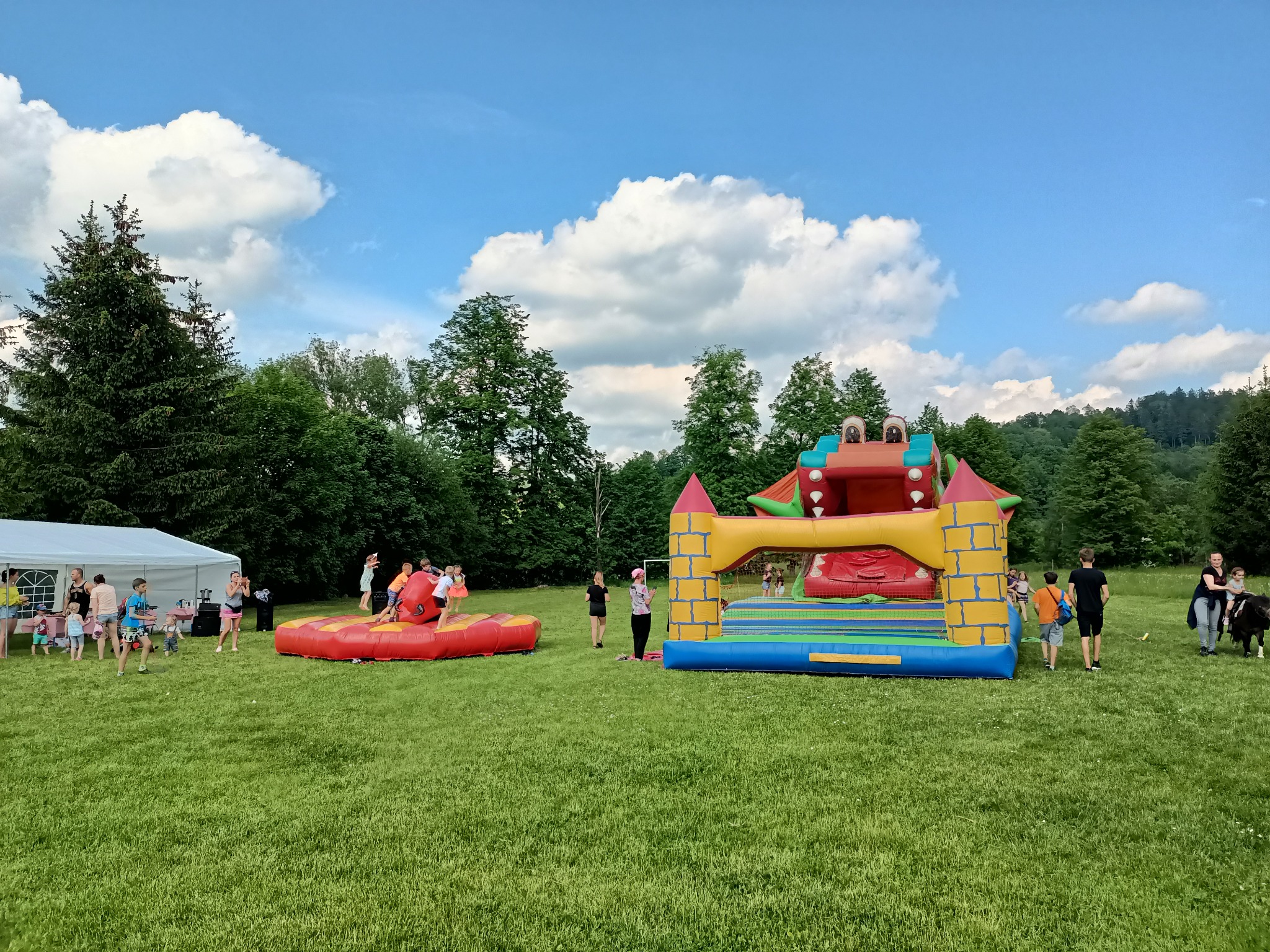 Dwa dmuchane zamki na zielonej trawie, dzieci bawiące się w pobliżu, biały namiot w tle, drzewa i błękitne niebo z chmurami.