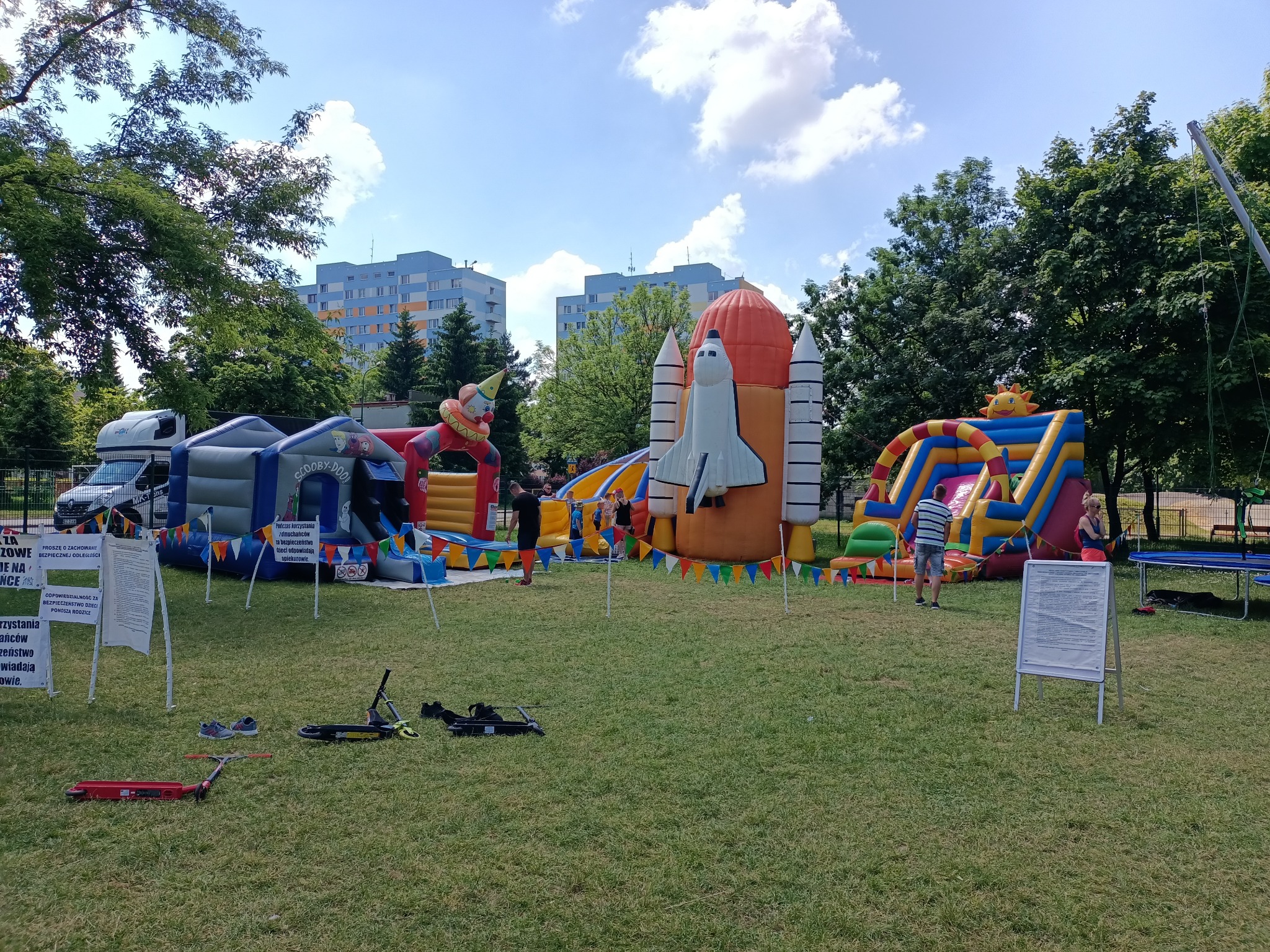 Rozległy plac zabaw z dmuchanymi atrakcjami, w tym rakieta kosmiczna i zjeżdżalnie, otoczony zielenią i blokami mieszkalnymi w tle, z widocznymi tablicami informacyjnymi i porzuconymi hulajnogami...