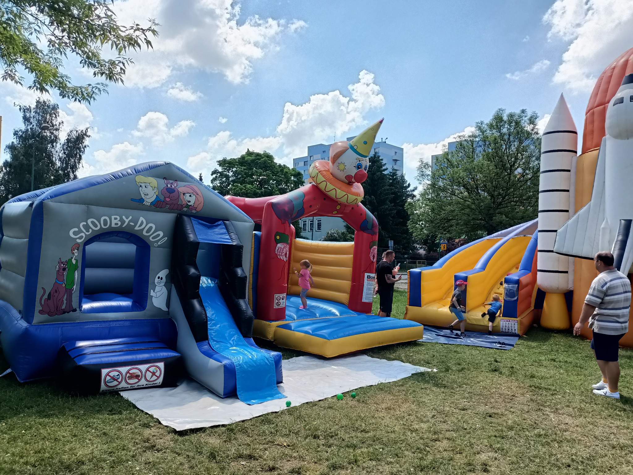 Kolorowe dmuchańce, w tym Scooby-Doo i rakieta kosmiczna, rozstawione na trawie w Świdnicy, z bawiącymi się dziećmi i nadzorującymi dorosłymi w słoneczny dzień.