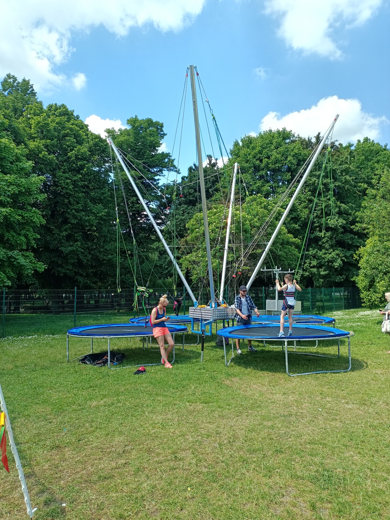 Rozstawione na trawie cztery stanowiska z trampolinami bungee, z których korzystają osoby dorosłe i dziecko, na tle drzew i błękitnego nieba.