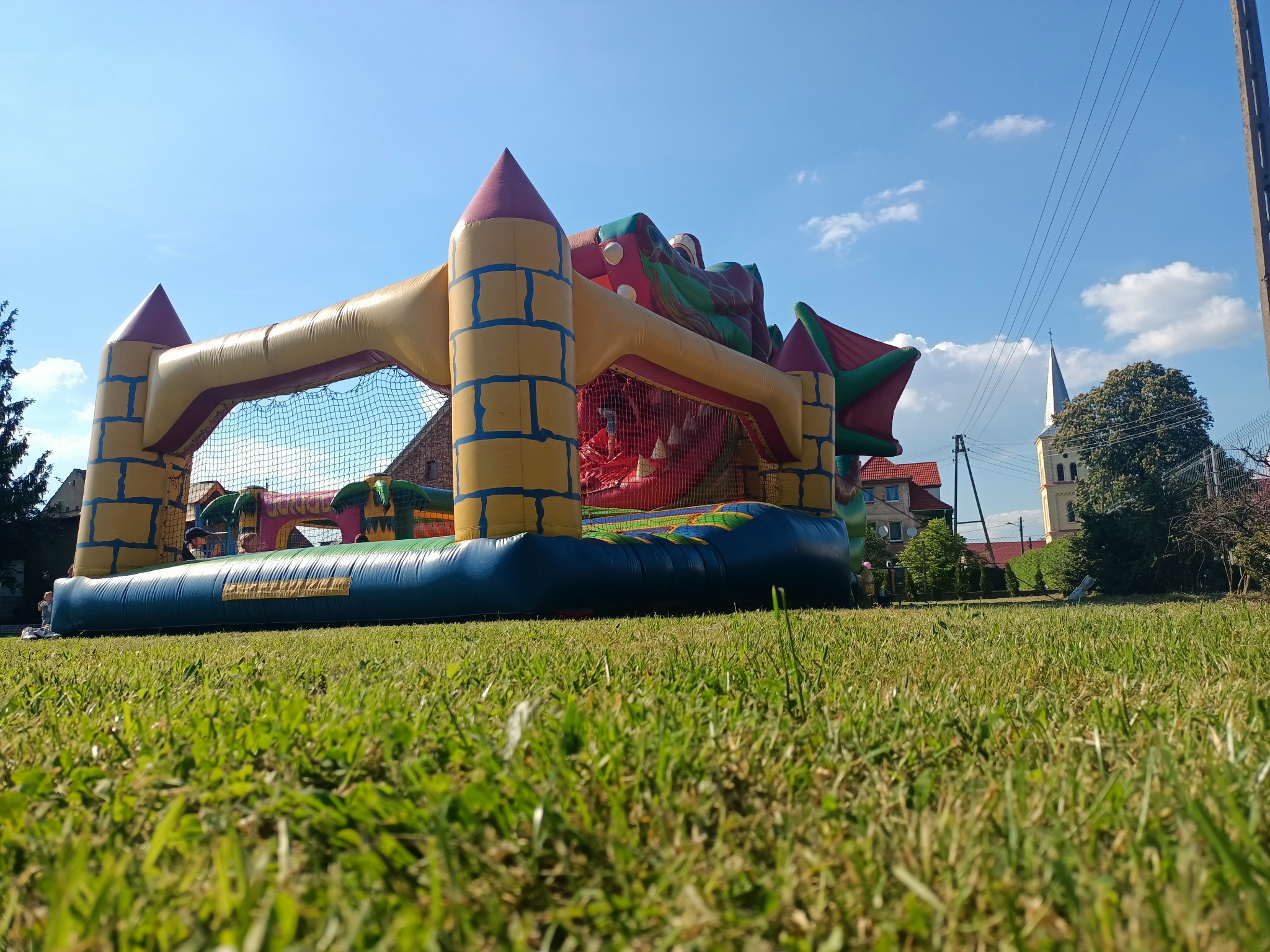 Duży, kolorowy dmuchany zamek na trawniku, widziany z perspektywy trawy, z kościołem w tle.