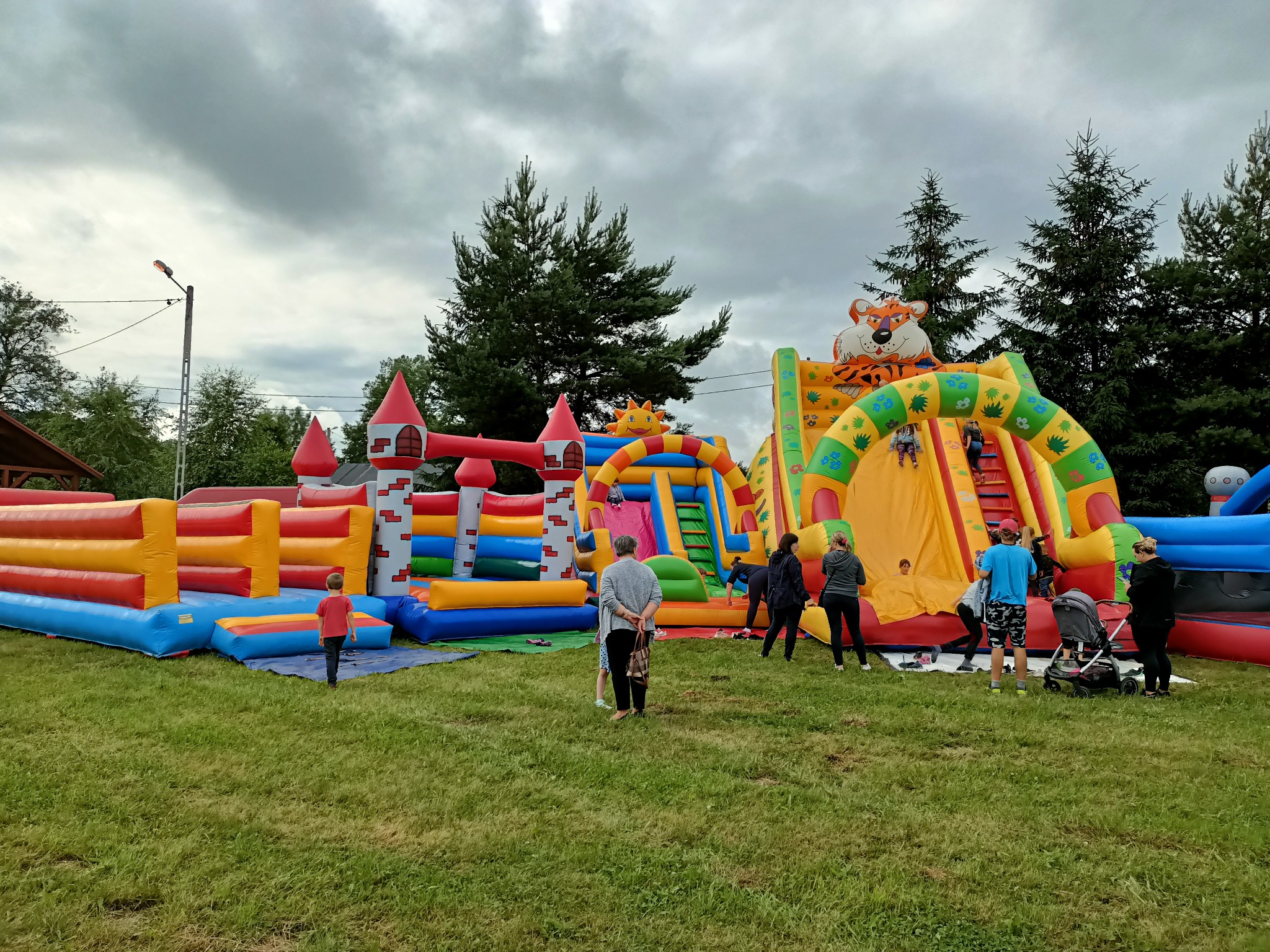 Kolorowe, dmuchane zamki i zjeżdżalnie z motywami zwierzęcymi na zielonej trawie, dzieci i dorośli w pobliżu, pochmurne niebo w tle.