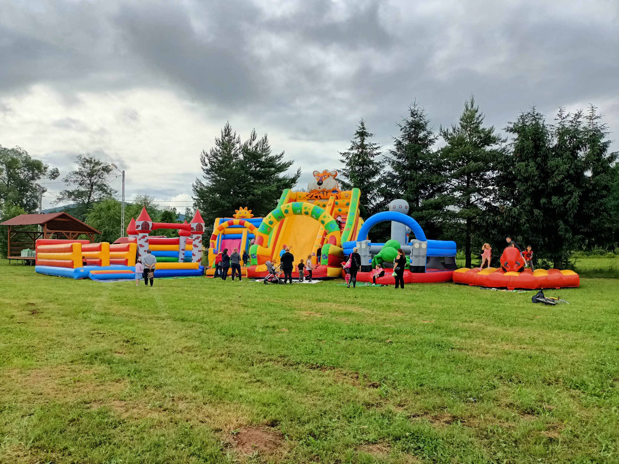 Rozległy plac zabaw na trawie z dmuchanymi zamkami, zjeżdżalniami i przeszkodami w kolorach tęczy, otoczony zielenią drzew i pochmurnym niebem, gdzie dzieci i dorośli korzystają z atrakcji.
