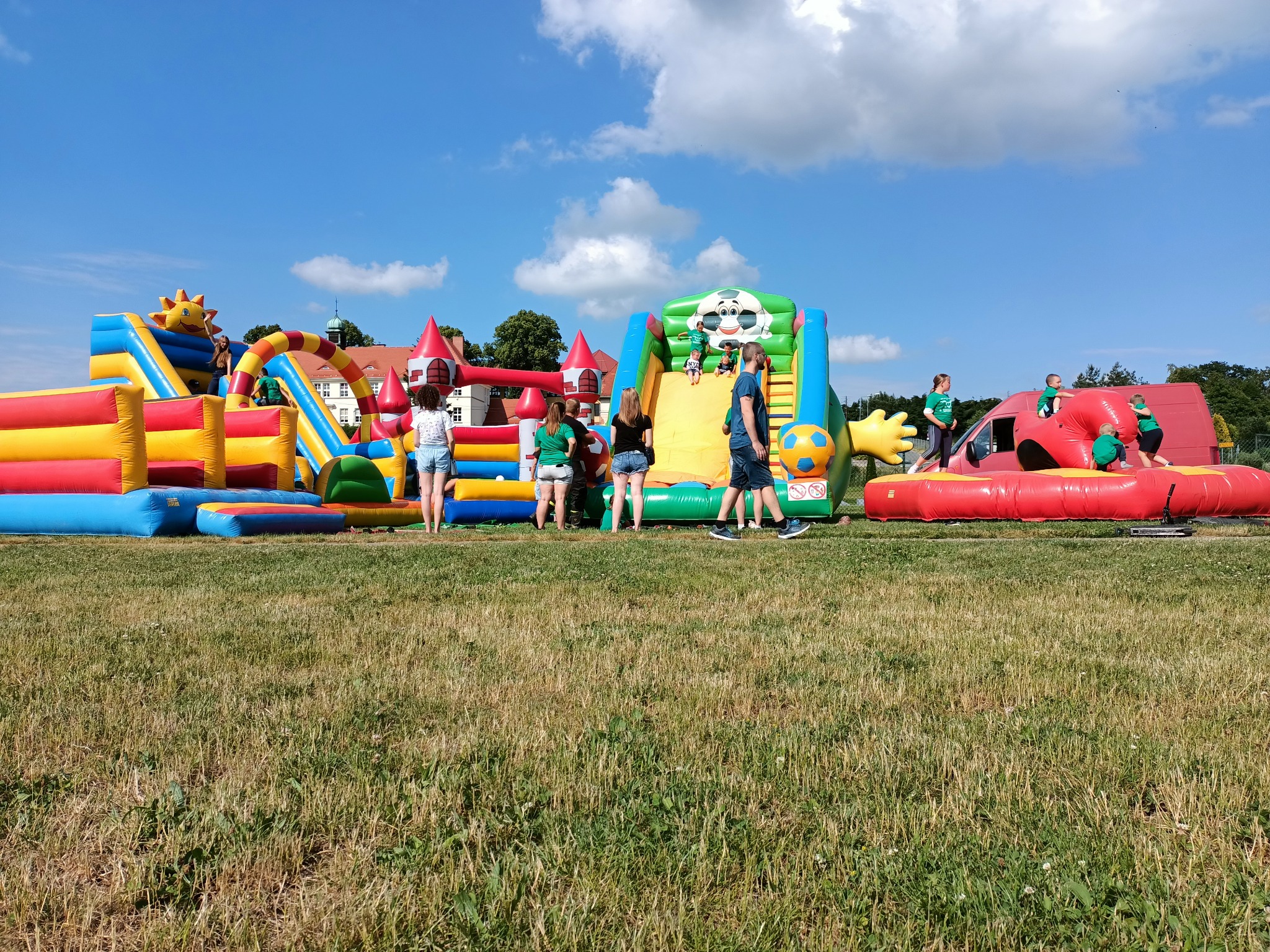 Kolorowy park dmuchanych zamków i zjeżdżalni na trawiastym terenie, dzieci i dorośli w tle, budynek z czerwonym dachem w oddali, błękitne niebo z chmurami.