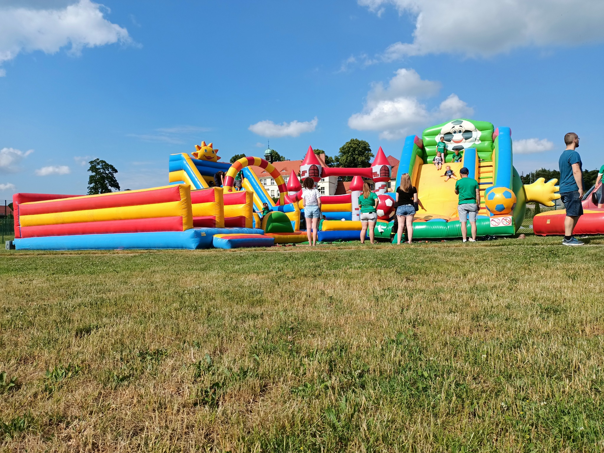 Kolorowy park dmuchanych atrakcji zjeżdżalni, zamków i przeszkód dla dzieci, rozstawiony na trawiastym terenie pod błękitnym niebem z białymi obłokami, z widocznymi osobami w tle.