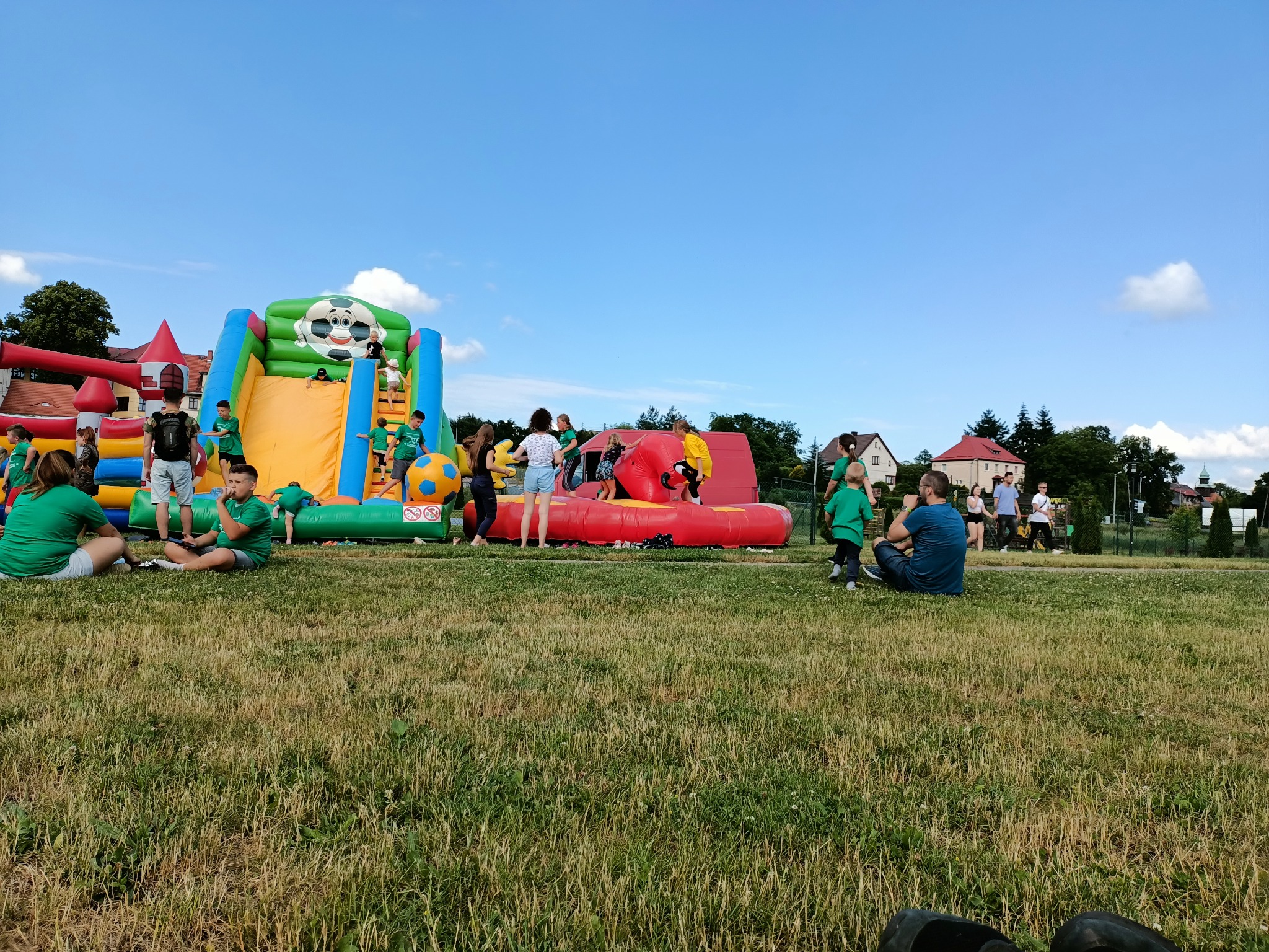 Ustawione na trawie kolorowe dmuchańce z motywem piłki nożnej, wokół bawiące się dzieci w zielonych koszulkach, w tle budynki mieszkalne pod błękitnym niebem.