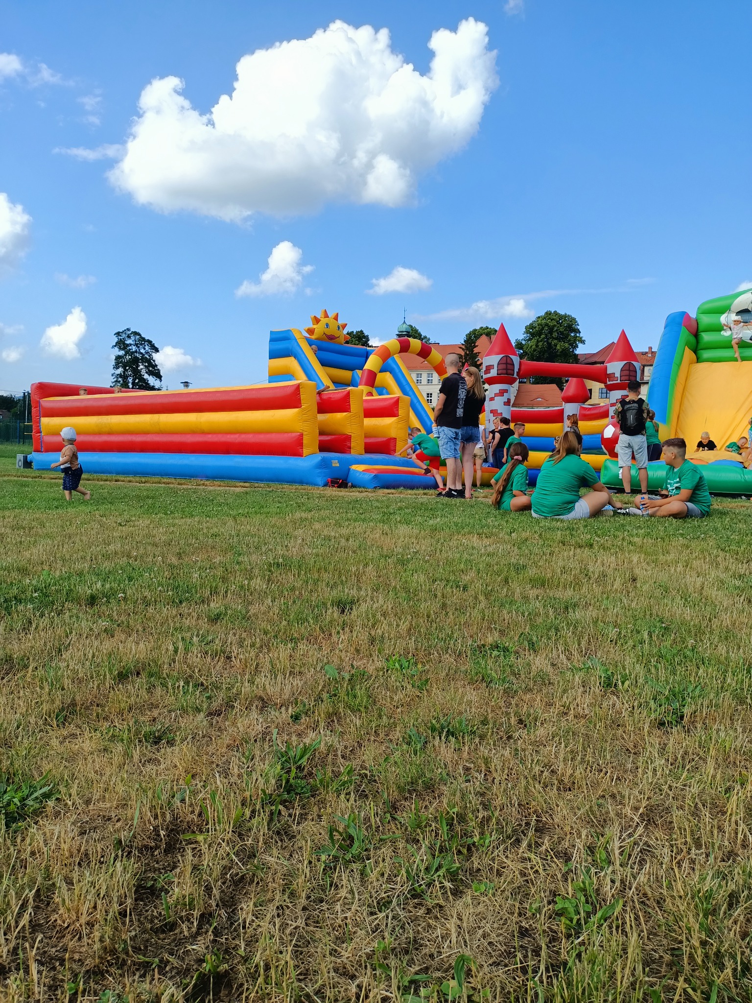 Kolorowe dmuchańce w kształcie zamku i zjeżdżalni na trawiastym terenie, dzieci w zielonych koszulkach odpoczywają obok, błękitne niebo z chmurami w tle.