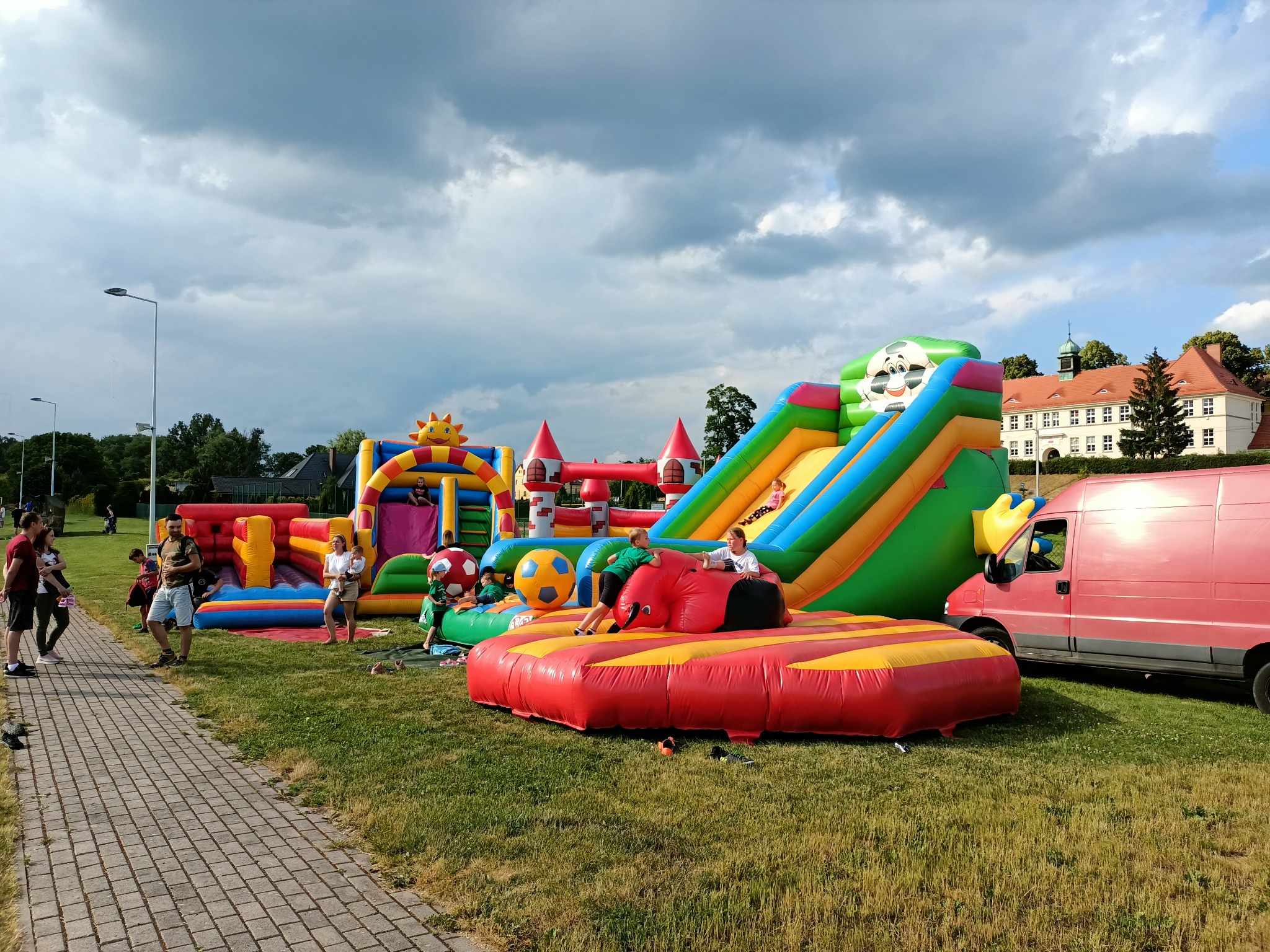 Kolorowe dmuchane zamki i zjeżdżalnie na zielonej trawie, dzieci bawią się na dmuchanych atrakcjach, w tle budynek z czerwonym dachem i pochmurne niebo.