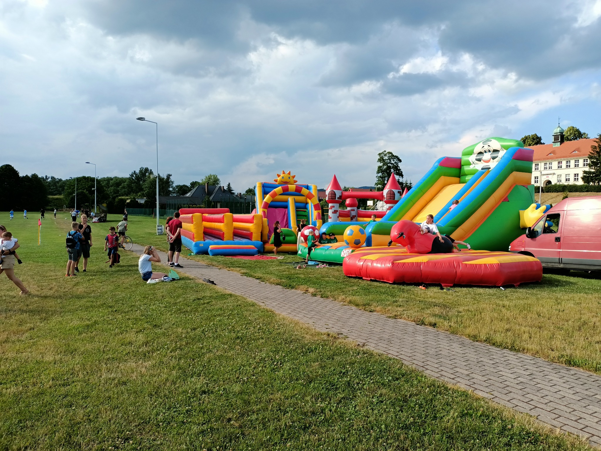 Kolorowe dmuchańce rozstawione na trawiastym terenie z bawiącymi się dziećmi, widoczny budynek w tle, pochmurne niebo.