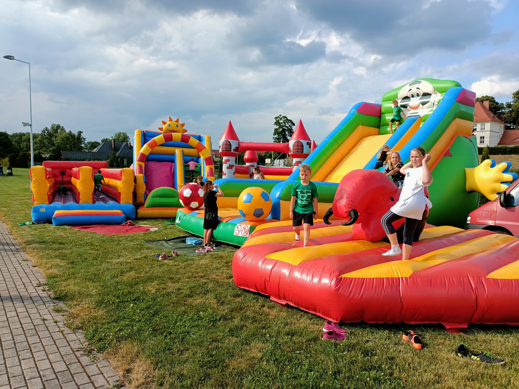 Dzieci bawiące się na rozłożonych, kolorowych dmuchańcach na trawniku w plenerze; w tle budynki i drzewa.