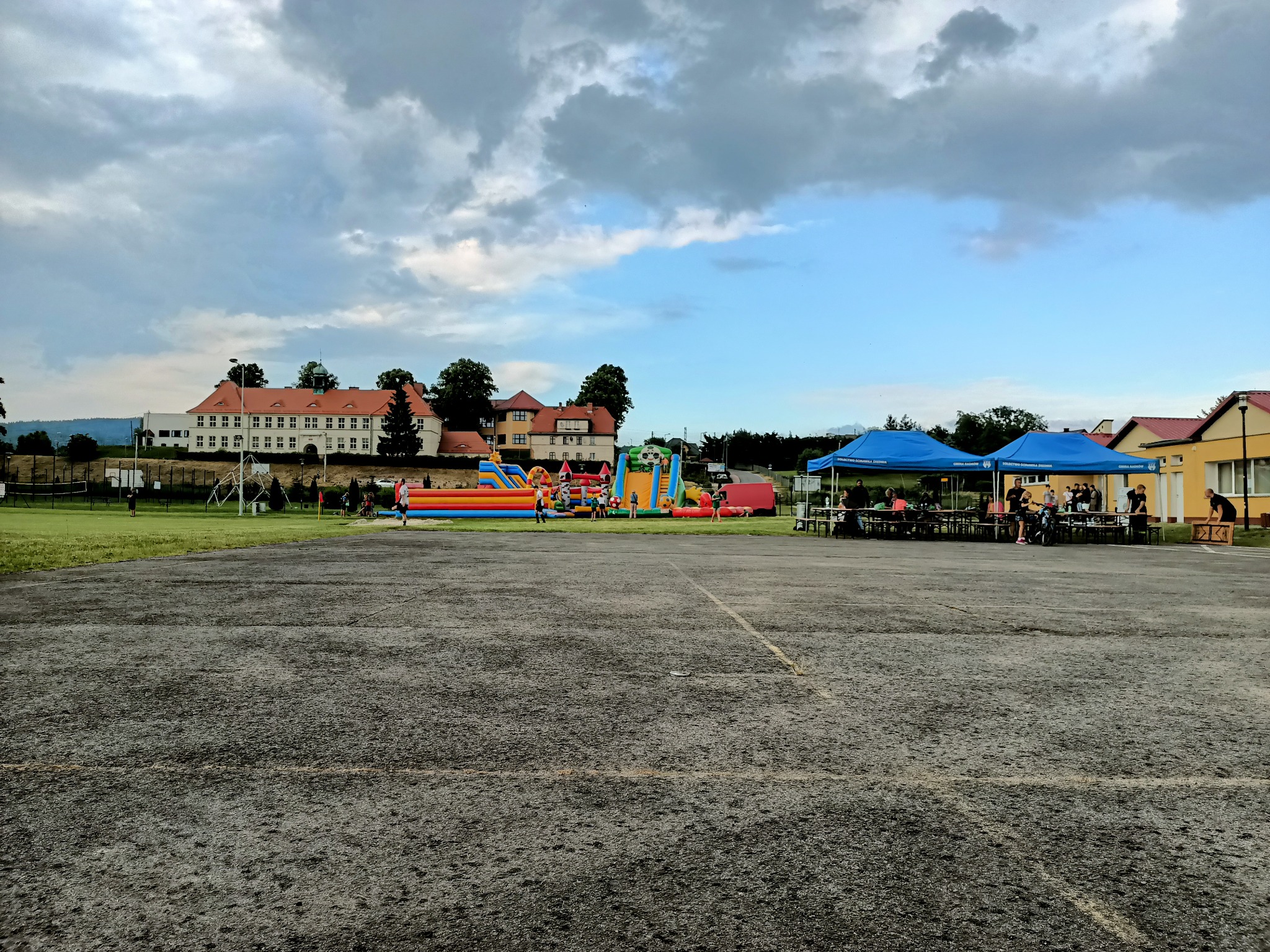 Rozległy plac z dmuchanymi zamkami i zjeżdżalniami w tle budynku, pod błękitnymi namiotami siedzą ludzie, pochmurne niebo.