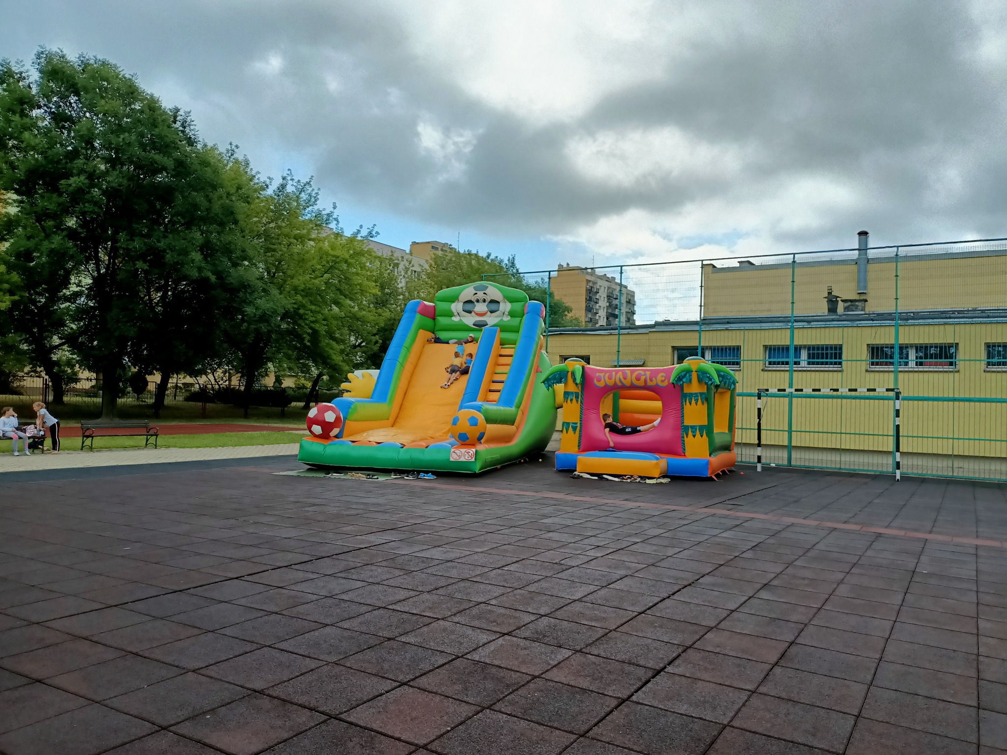 Dwa dmuchane zamki na placu zabaw: duża zjeżdżalnia w kształcie boiska piłkarskiego i mniejszy domek z napisem 'Jungle'. Dzieci bawią się na zjeżdżalni. W tle budynek szkolny i drzewa.