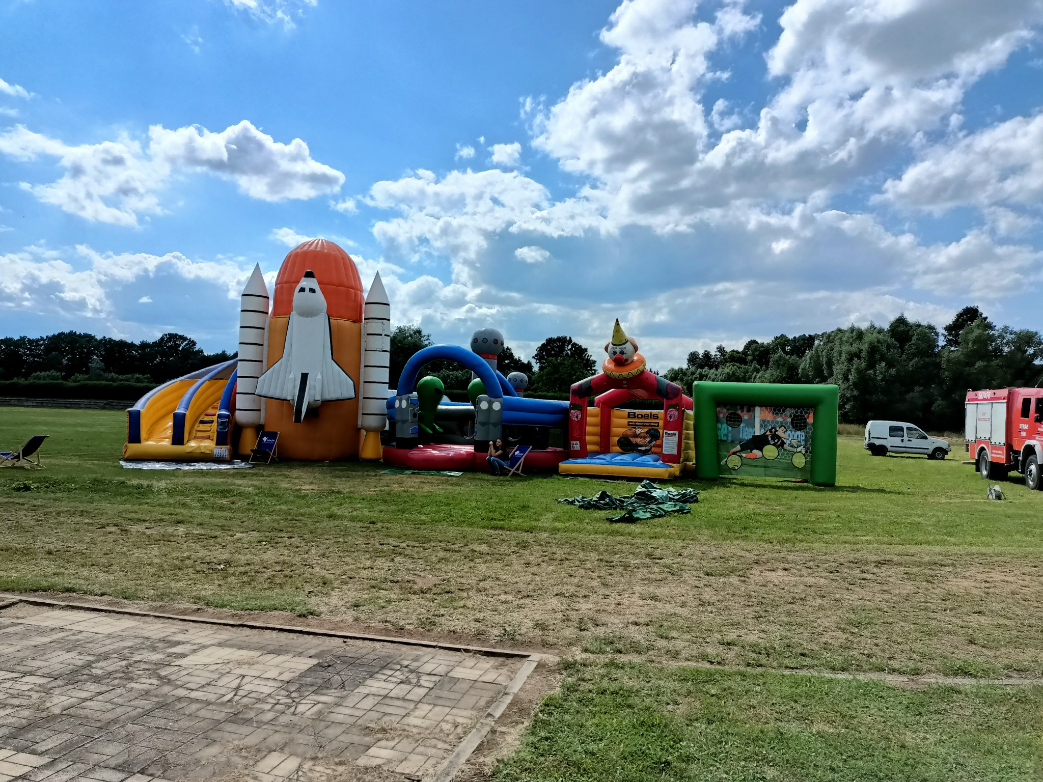 Kompleks dmuchańców na trawie: rakieta kosmiczna, tor przeszkód, klaun z basenem, bramka piłkarska, w tle samochód strażacki i dostawczy.