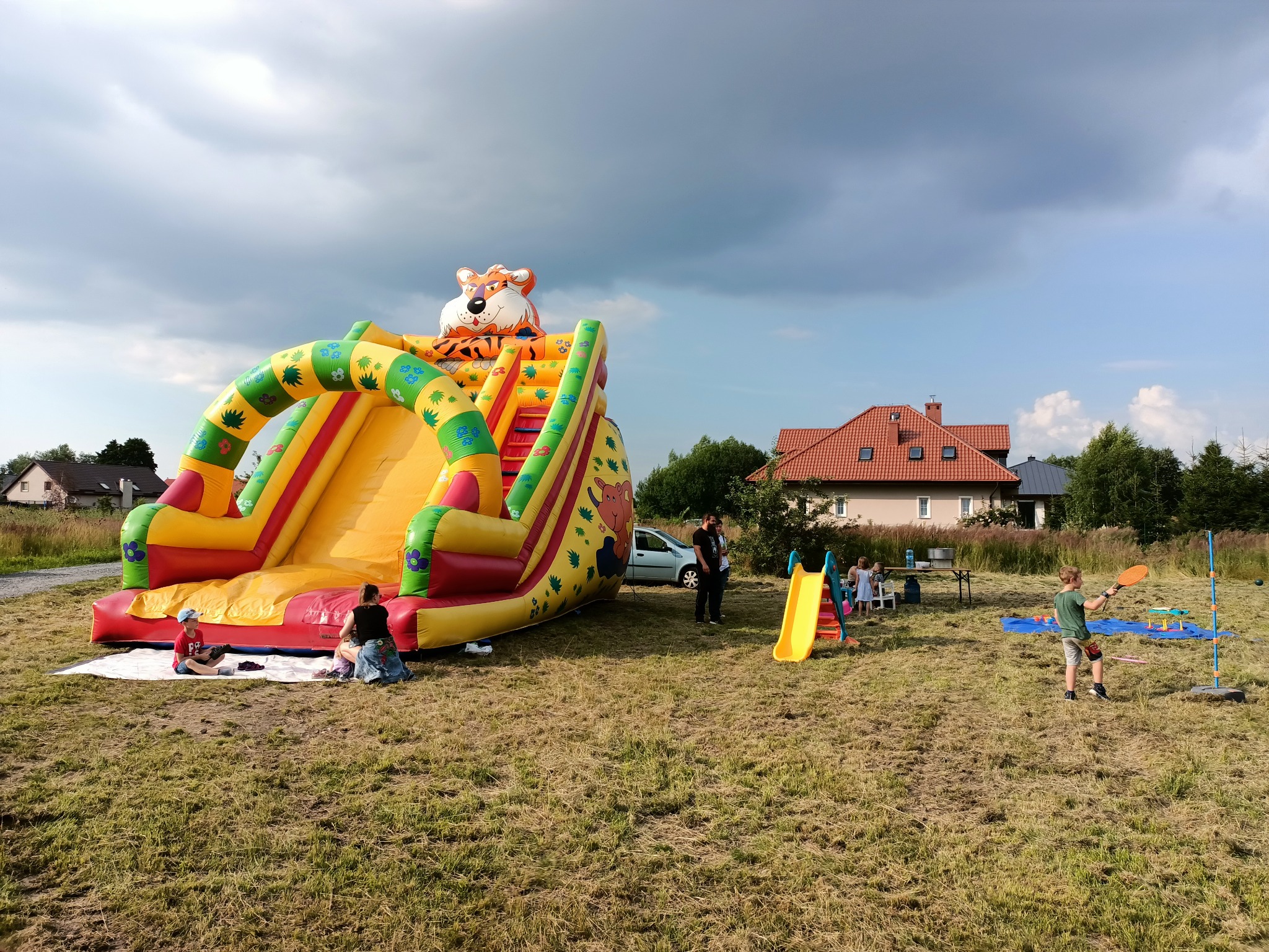 Duża, dmuchana zjeżdżalnia z motywem tygrysa na szczycie, ustawiona na trawniku podczas imprezy plenerowej, w tle domy i pochmurne niebo.