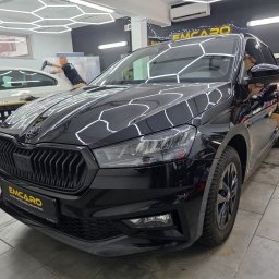 EMCARO Auto Detailing & Wrapping - Błyszcząca, czarna Skoda Fabia po detailingu w studiu EMCARO. Widoczne odbicia światła na lakierze, w tle pracownik aplikujący folię na inny pojazd.