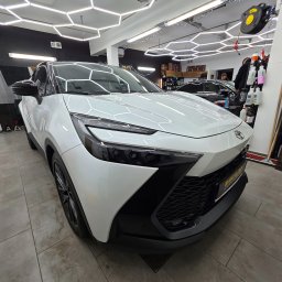 EMCARO Auto Detailing & Wrapping - Biały samochód Toyota C-HR po detailingu w jasnym wnętrzu myjni, z widocznymi lampami LED i akcesoriami do pielęgnacji aut w tle. Błyszcząca karoseria.