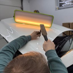 EMCARO Auto Detailing & Wrapping - Usuwanie wgniecenia na białej masce samochodu przy użyciu lampy inspekcyjnej i narzędzia PDR. Widoczne ręce pracownika w trakcie naprawy.
