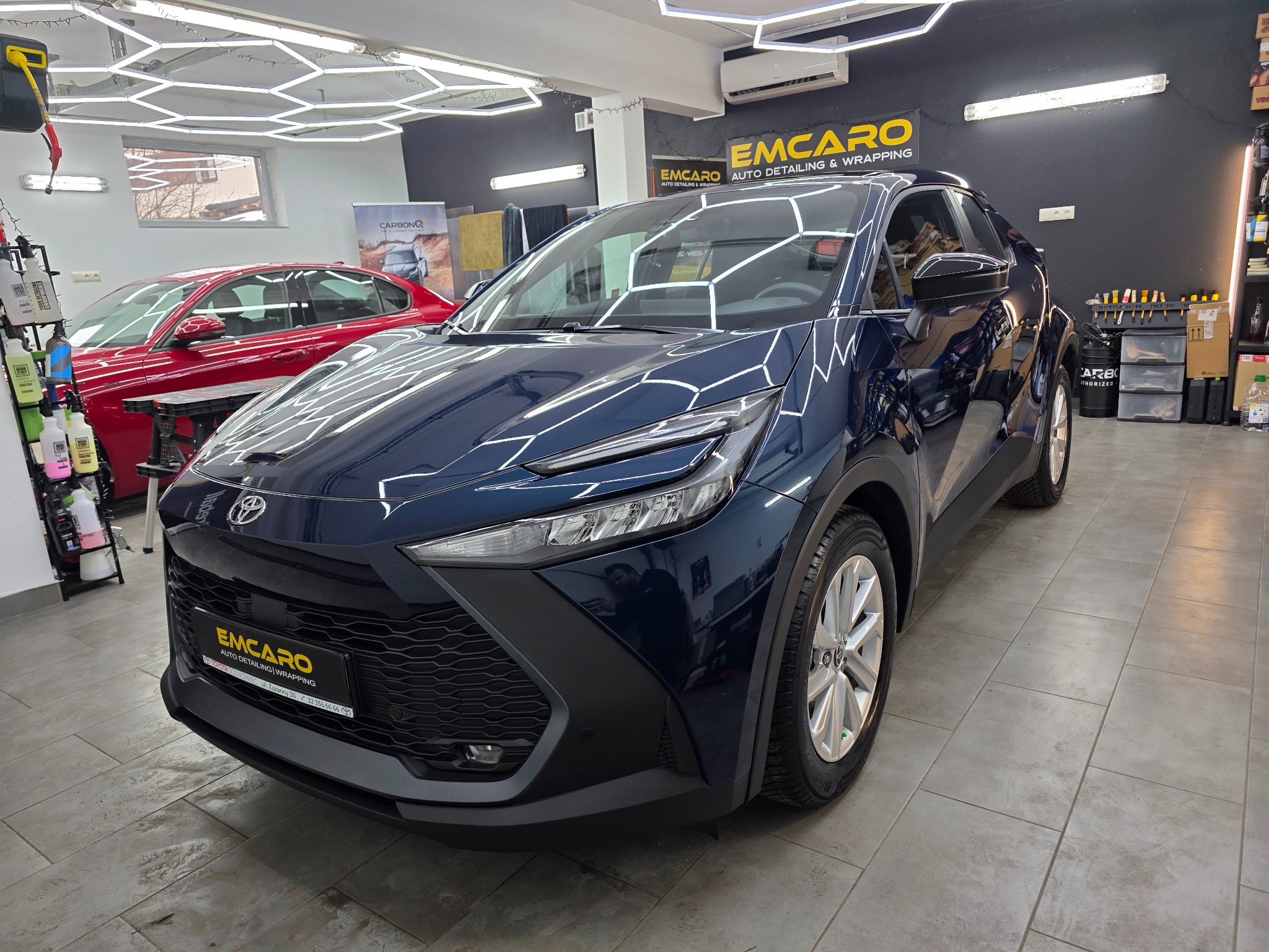 Błyszczący, granatowy samochód po detailingu w studiu. Widoczne logo firmy detailingowej na pojeździe i w tle. Oświetlenie LED podkreśla gładkość lakieru.