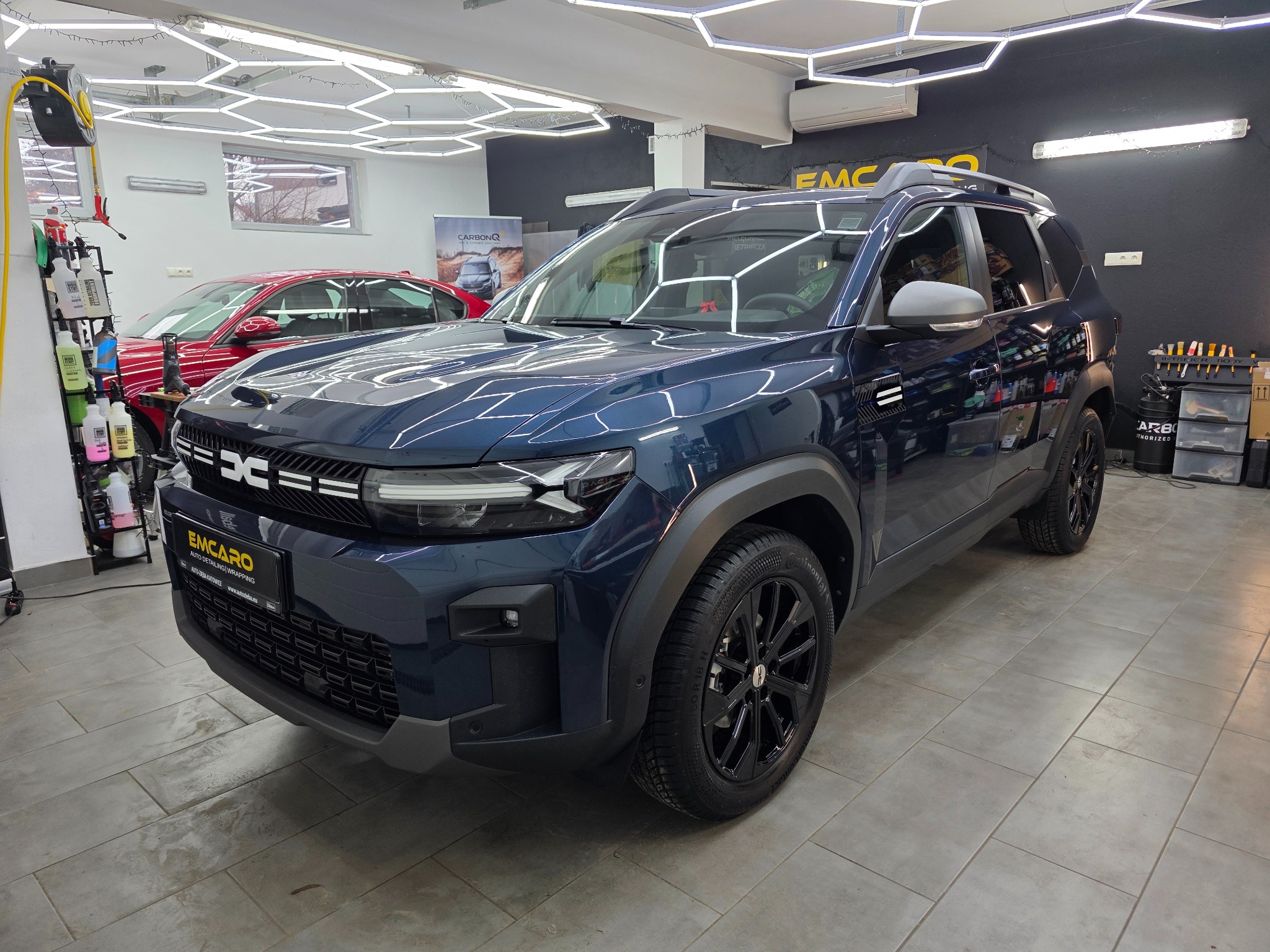 Błyszczący, granatowy SUV Dacia po detailingu w studiu EMCARO. Widoczne narzędzia i kosmetyki. Oświetlenie LED w kształcie plastra miodu. Czyste felgi i karoseria.