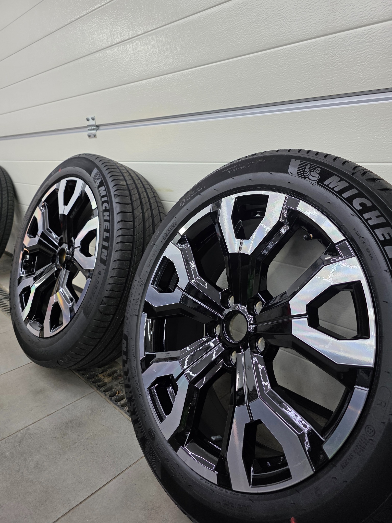 Dwa czarne, błyszczące koła samochodowe Michelin z aluminiowymi felgami o unikalnym wzorze, ustawione na szarym tle, w warsztacie.