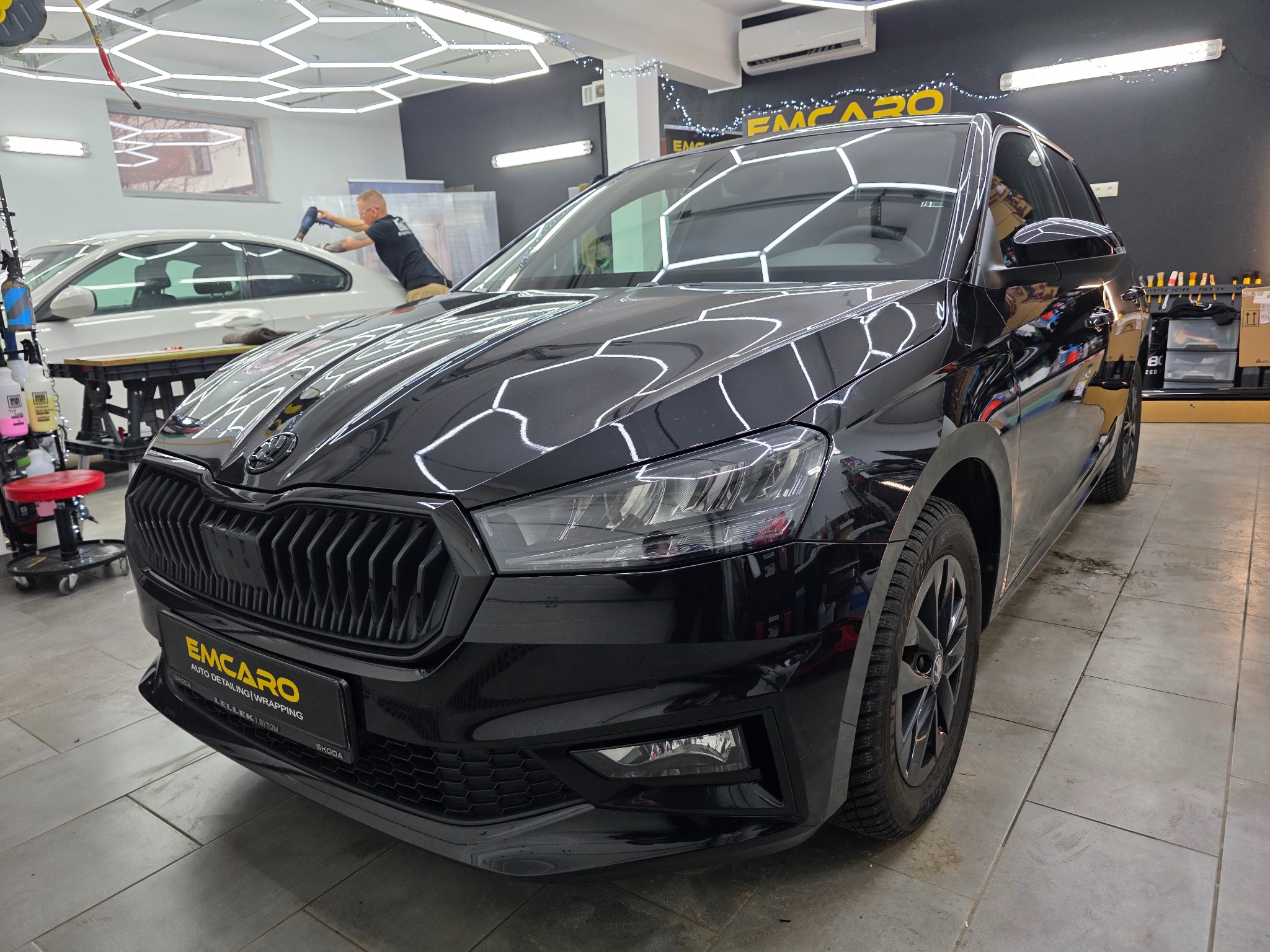 Błyszcząca, czarna Skoda Fabia po detailingu w studiu EMCARO. Widoczne odbicia światła na lakierze, w tle pracownik aplikujący folię na inny pojazd.