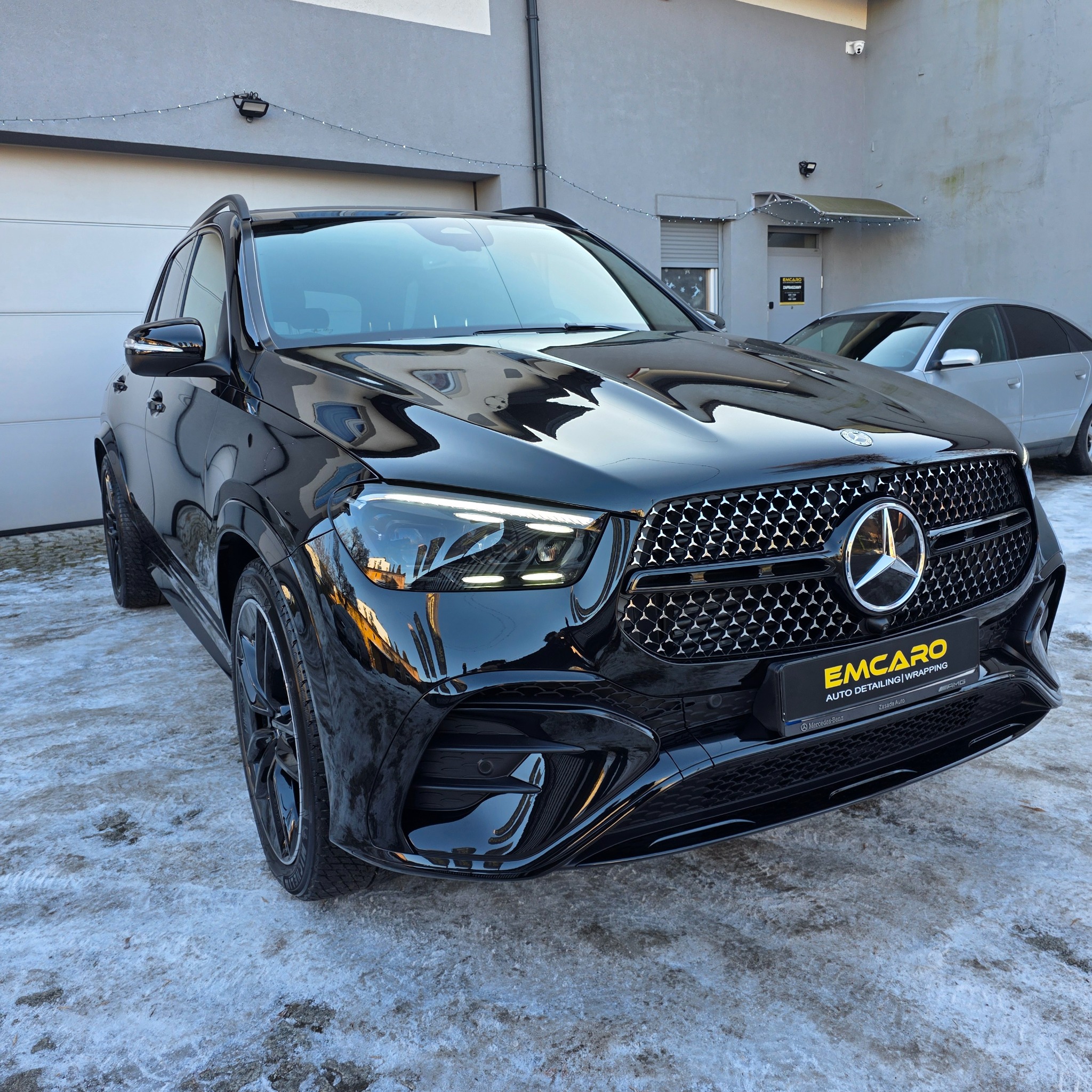 Błyszczący, czarny Mercedes GLE po detailingu, z widocznym logo firmy detailingowej na tle zimowego otoczenia. Reflektory włączone, podkreślają efekt.