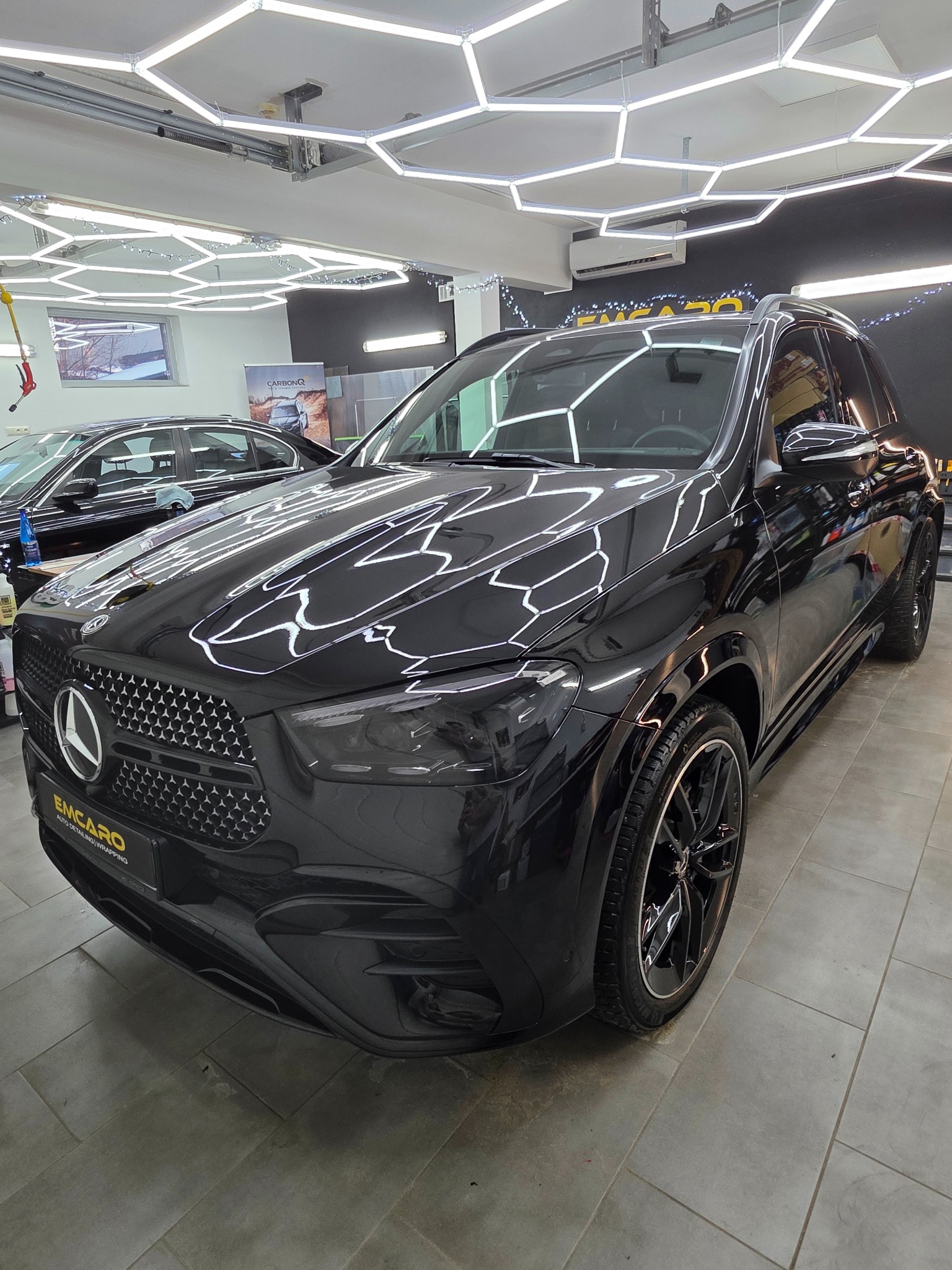 Błyszczący, czarny Mercedes GLE po detailingu, widoczny z przodu w dobrze oświetlonym pomieszczeniu. Refleksy świetlne podkreślają gładkość lakieru. Widoczne logo firmy.