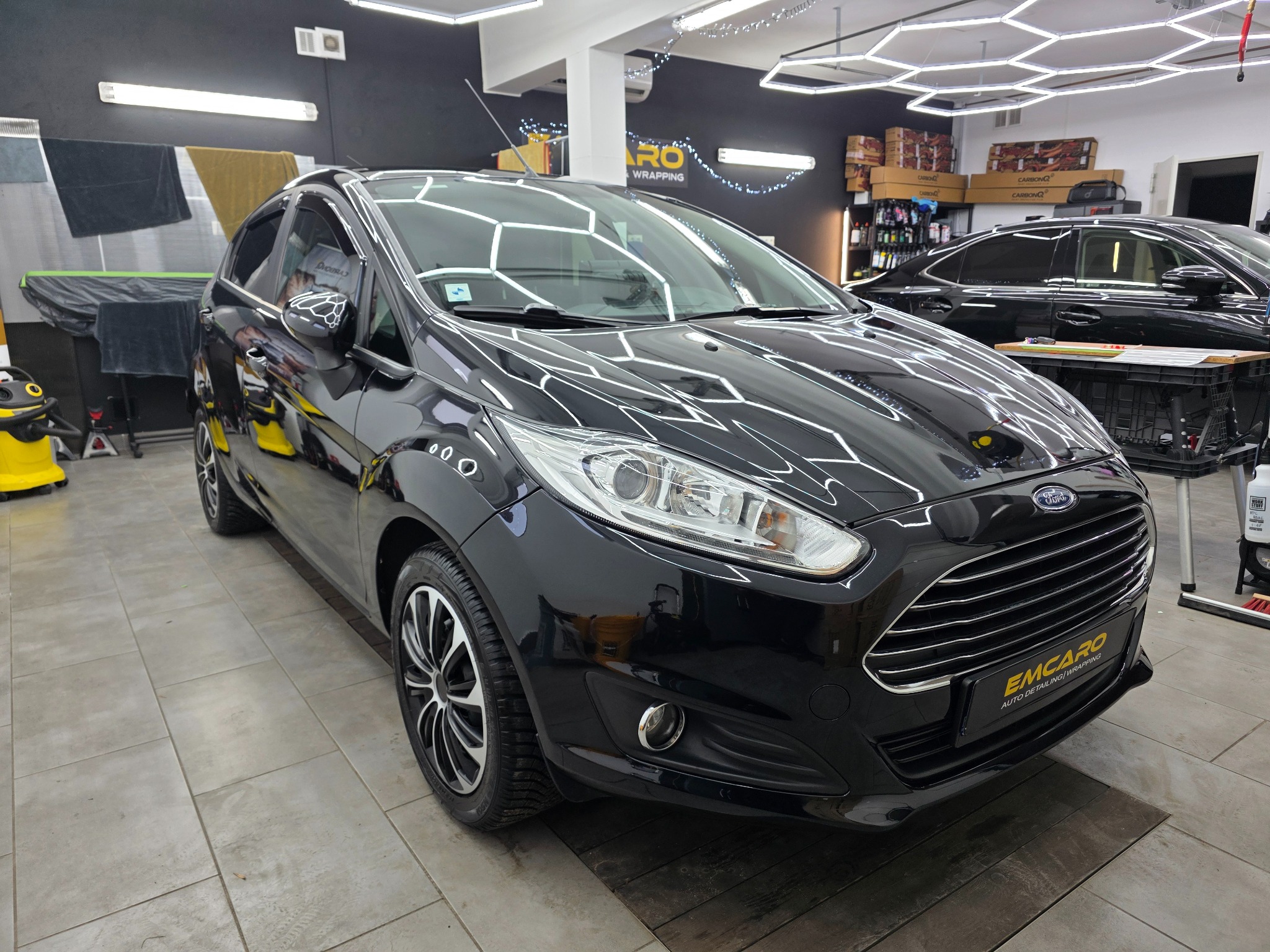 Błyszczący, czarny Ford Fiesta po detailingu w studiu. Widoczne oświetlenie heksagonalne, drugi samochód w tle i logo 'EMCARO Auto Detailing / Wrapping'.