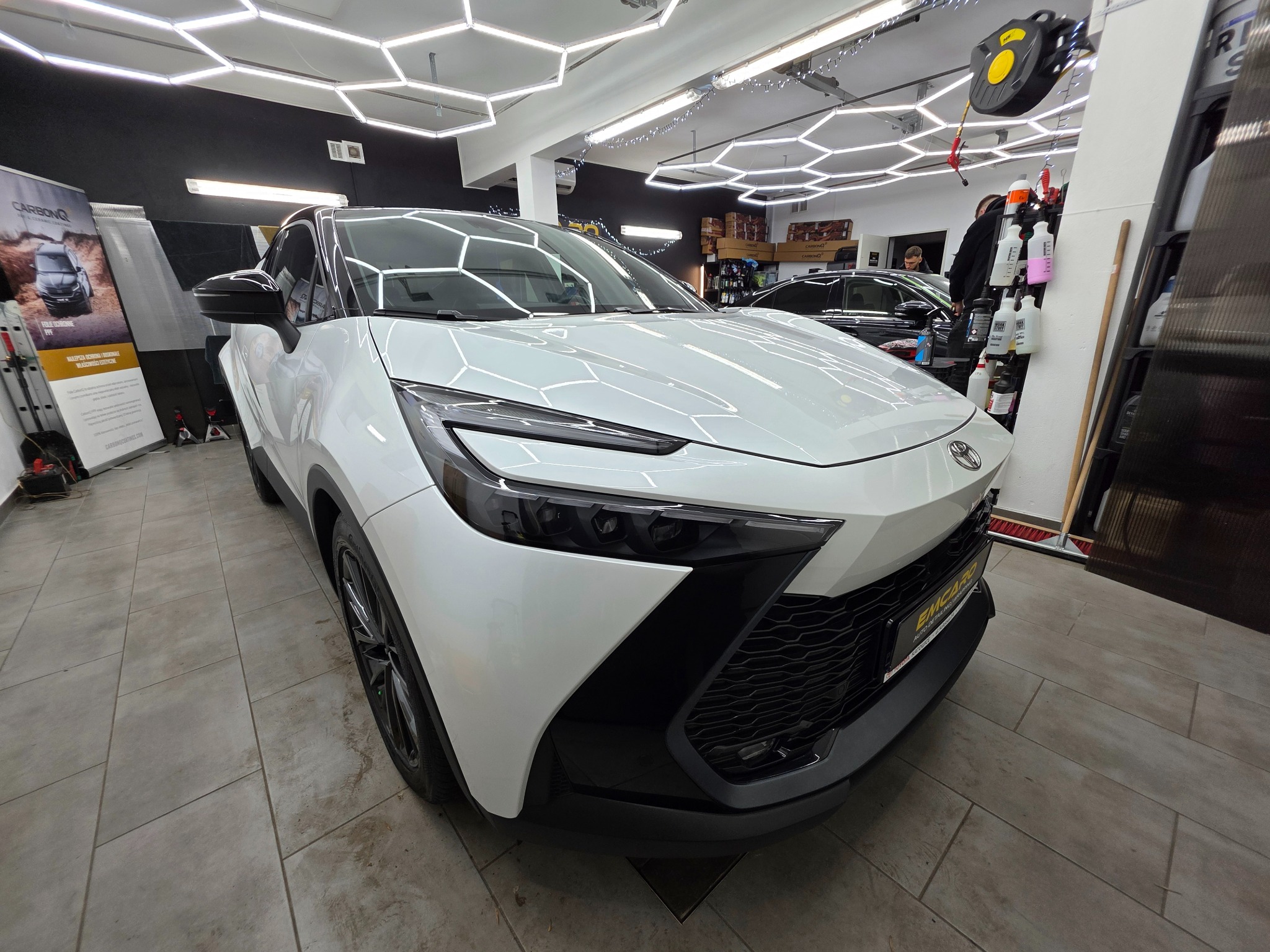 Biały samochód Toyota C-HR po detailingu w jasnym wnętrzu myjni, z widocznymi lampami LED i akcesoriami do pielęgnacji aut w tle. Błyszcząca karoseria.