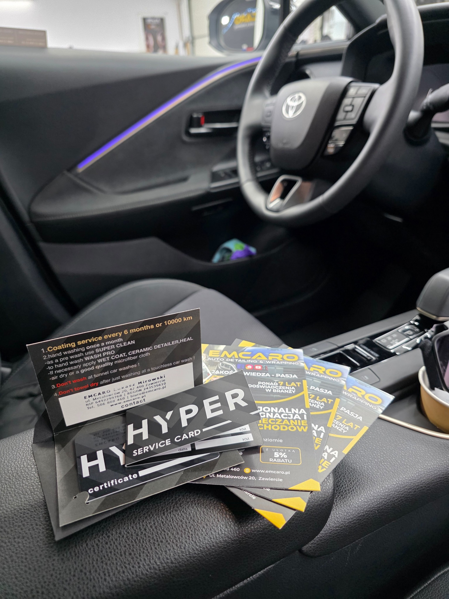 Wnętrze Toyoty z kartami detailingowymi EMCARO, prezentującymi usługi: powłoka ochronna, mycie ręczne, wrapping. Widoczne logo firmy i hasła: wiedza, pasja, doświadczenie.