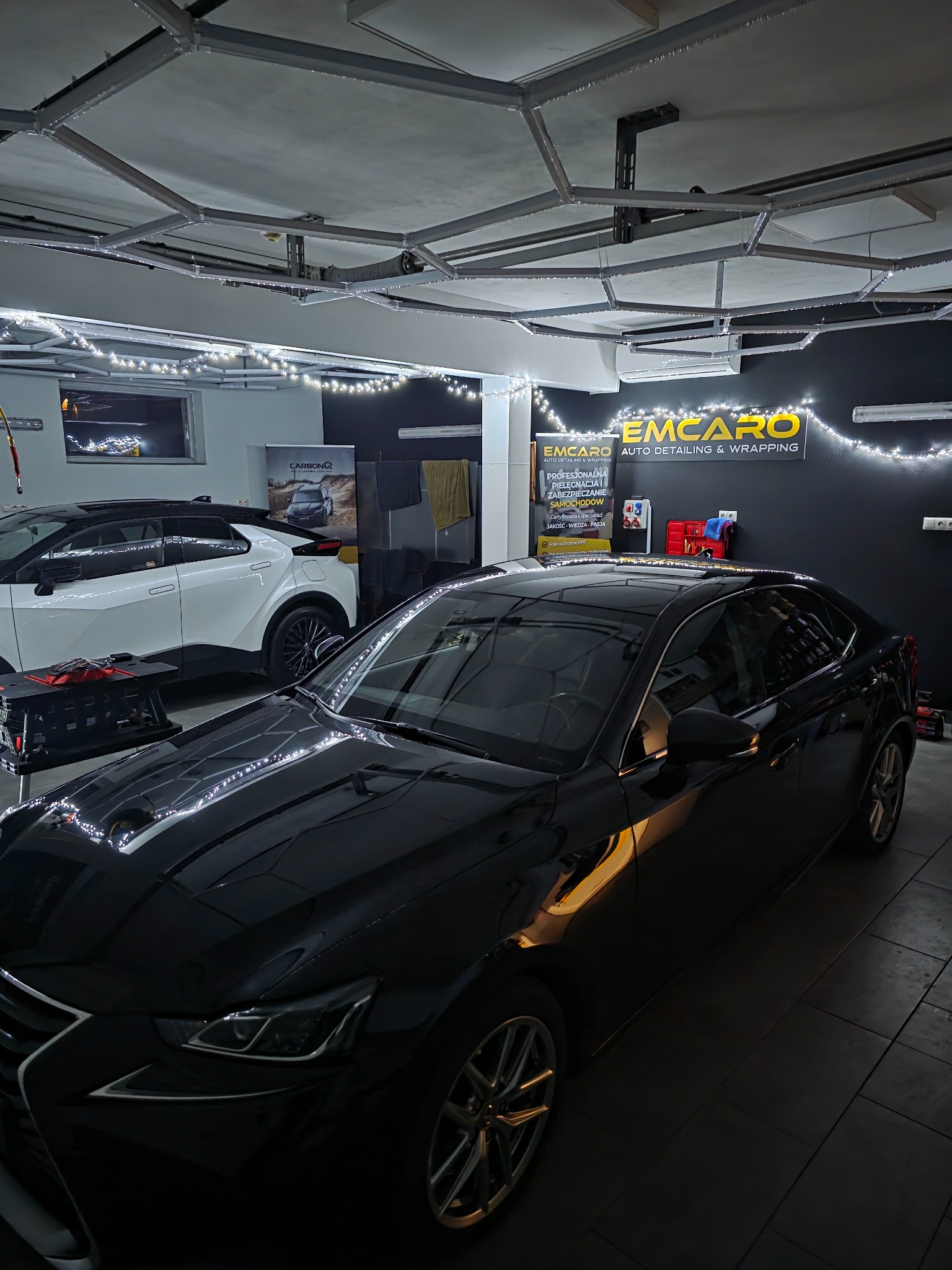 Błyszczący, czarny Lexus w studiu auto detailingu z dekoracyjnym oświetleniem. W tle biały samochód i logo firmy EMCARO. Widoczne narzędzia i akcesoria.