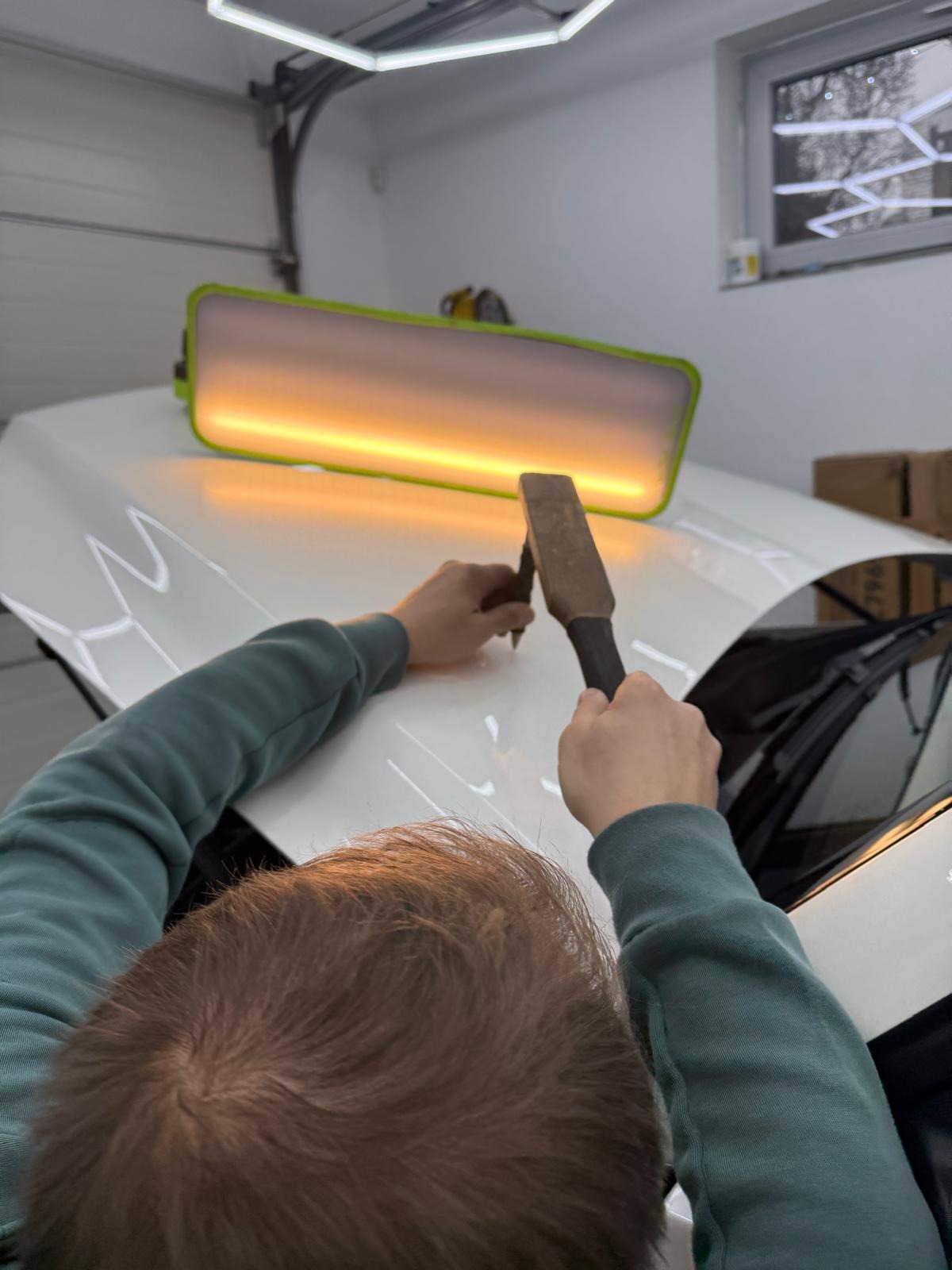 Usuwanie wgniecenia na białej masce samochodu przy użyciu lampy inspekcyjnej i narzędzia PDR. Widoczne ręce pracownika w trakcie naprawy.