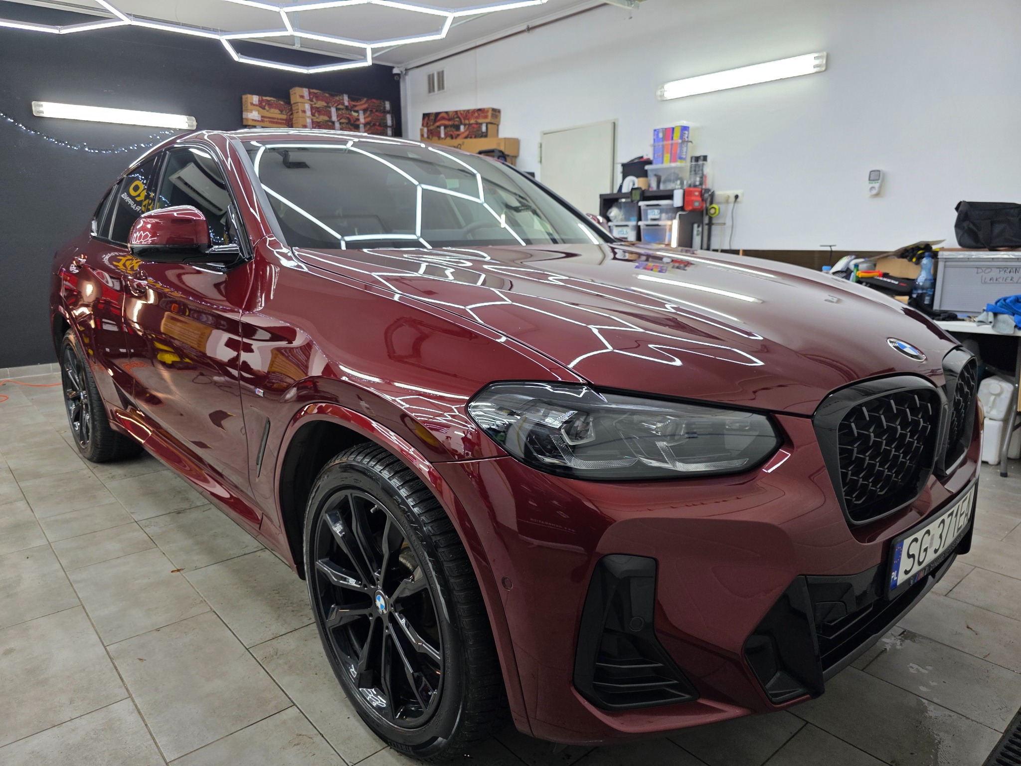Błyszczący, bordowy BMW X4 po detailingu w studiu, widoczne odbicia światła na lakierze, czarne felgi, oświetlenie LED na suficie. Tablica rejestracyjna widoczna.