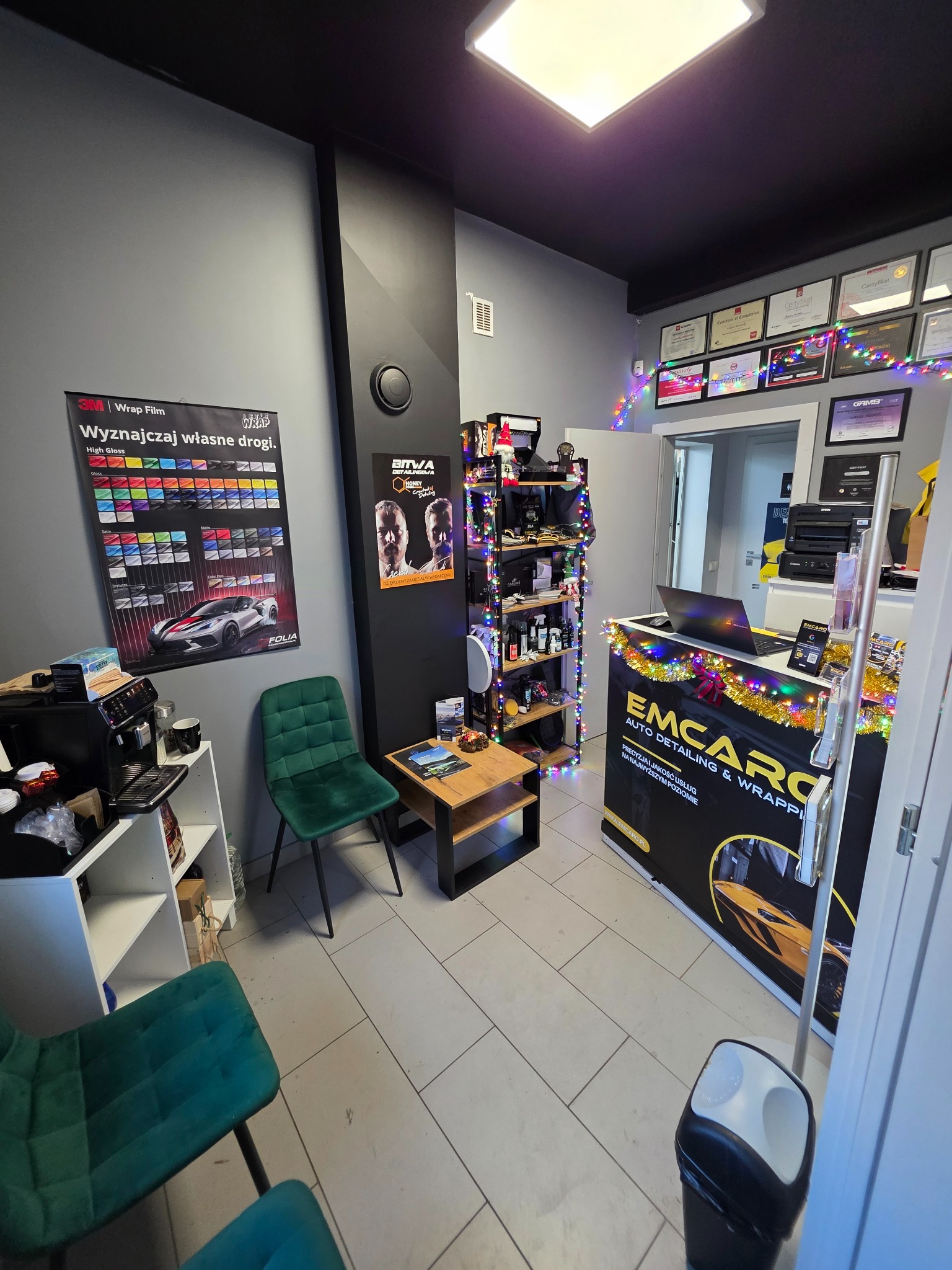 Wnętrze studia auto detailingu z recepcją, półkami z produktami, plakatami folii do oklejania aut i certyfikatami na ścianie. Świąteczny wystrój dodaje klimatu.