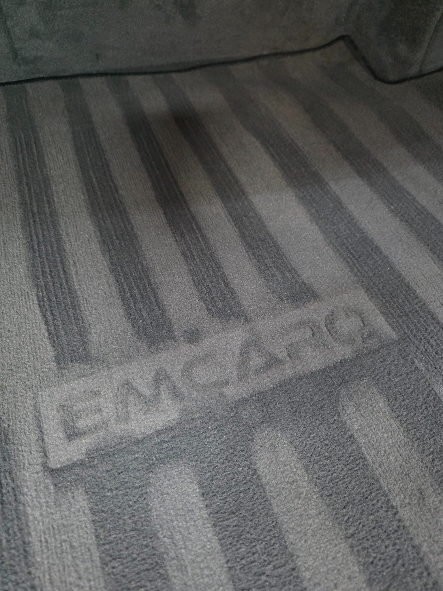 Szczegółowe ujęcie wyczyszczonej, szarej tapicerki samochodowej z widocznym wzorem w paski i napisem 'EMCARO', ukazujące efekt prania.