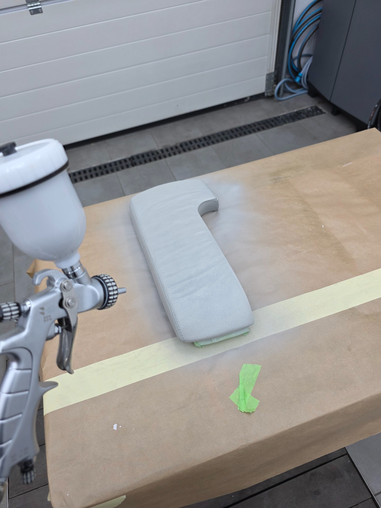 Tapicerowany podłokietnik samochodowy w trakcie renowacji, malowany pistoletem natryskowym na stole z papierem maskującym. Widoczny fragment garażu.