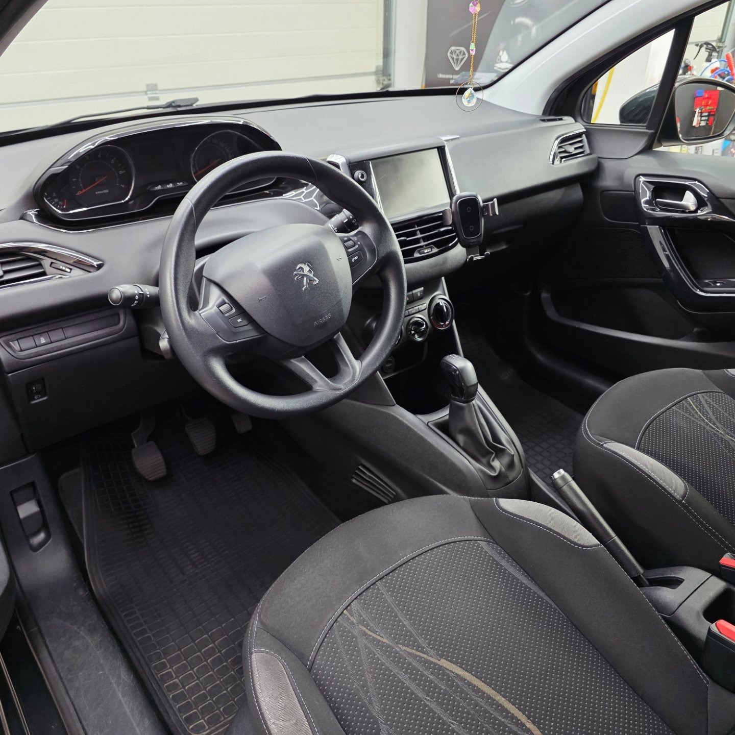 Wnętrze samochodu Peugeot 208 z czarną tapicerką, widok na deskę rozdzielczą, kierownicę z logo, fotele z szarymi wstawkami i gumowe dywaniki.