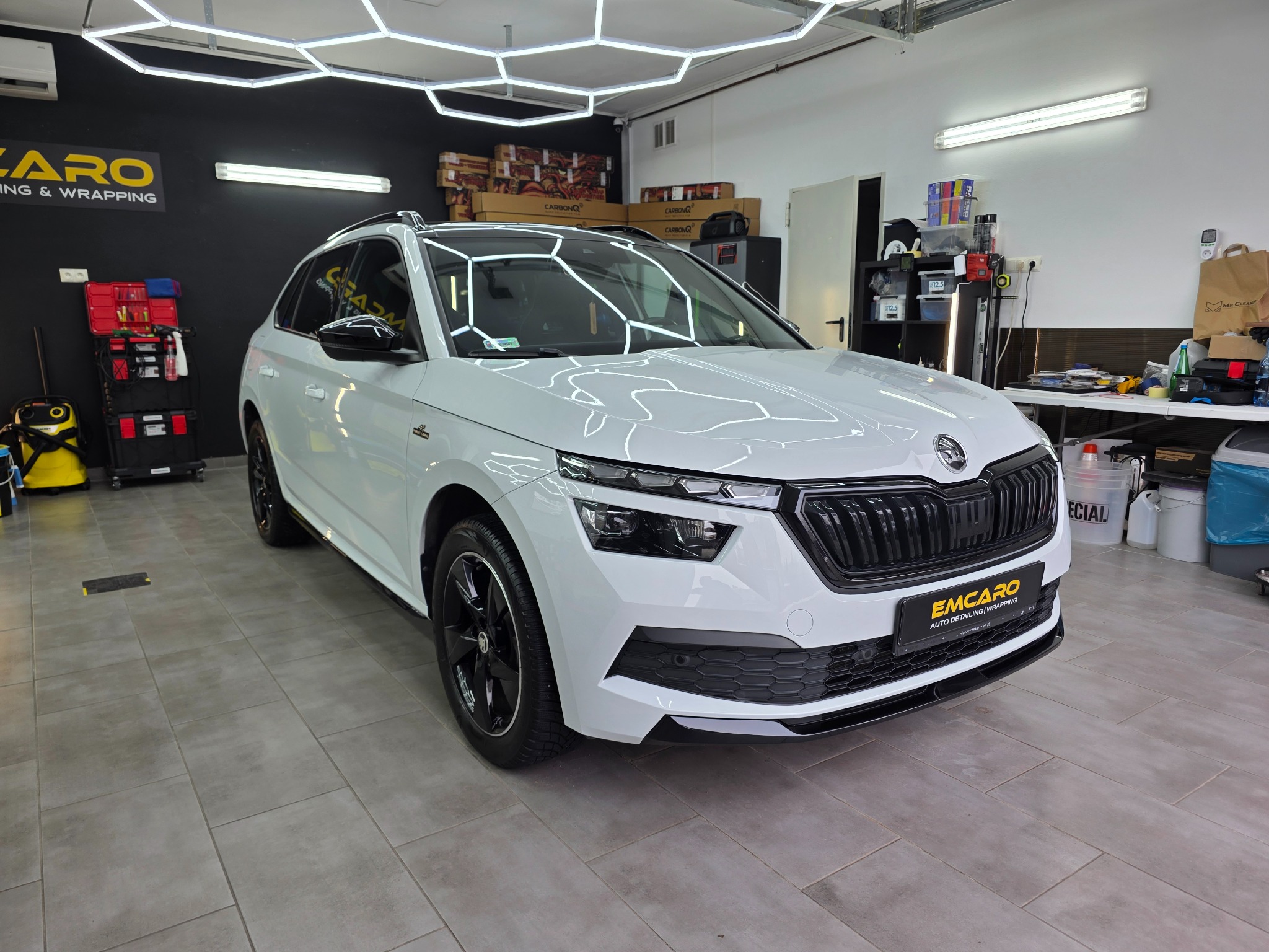 Biała Skoda Kamiq po detailingu w studiu, z widocznym logo EMCARO na tablicy rejestracyjnej. Oświetlenie sufitowe w kształcie plastra miodu. Błyszcząca karoseria.