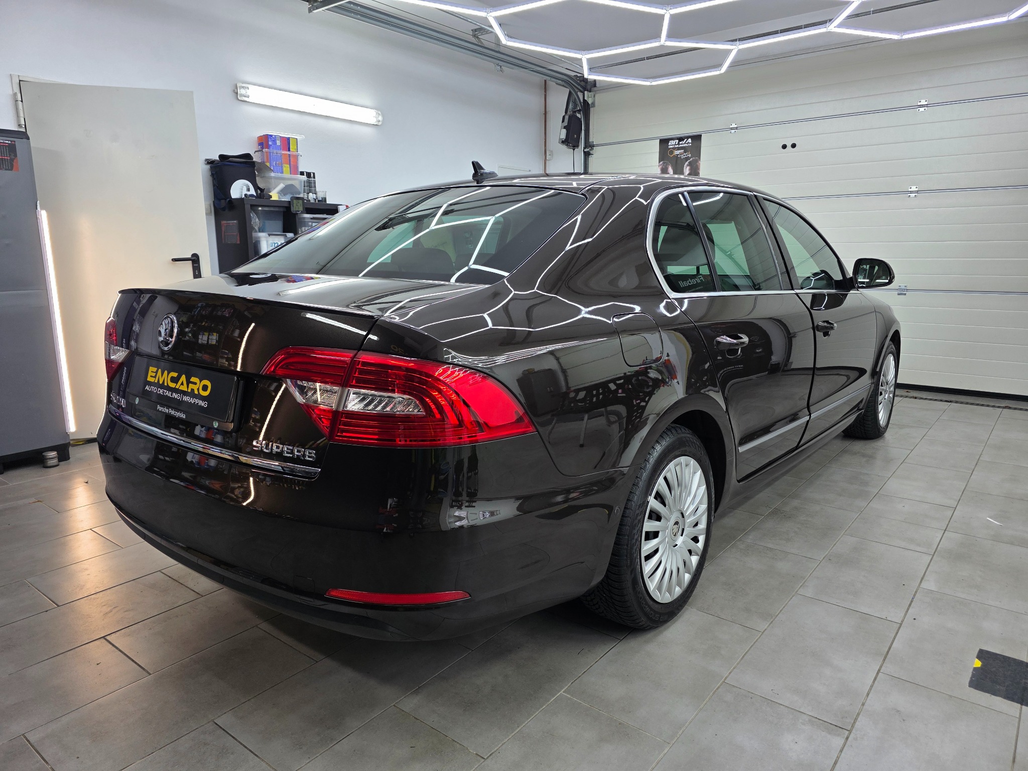 Błyszczący, ciemny sedan Skoda Superb po detailingu, zaparkowany w jasnym garażu z oświetleniem LED. Widoczny znak firmy detailingowej na tylnej klapie.
